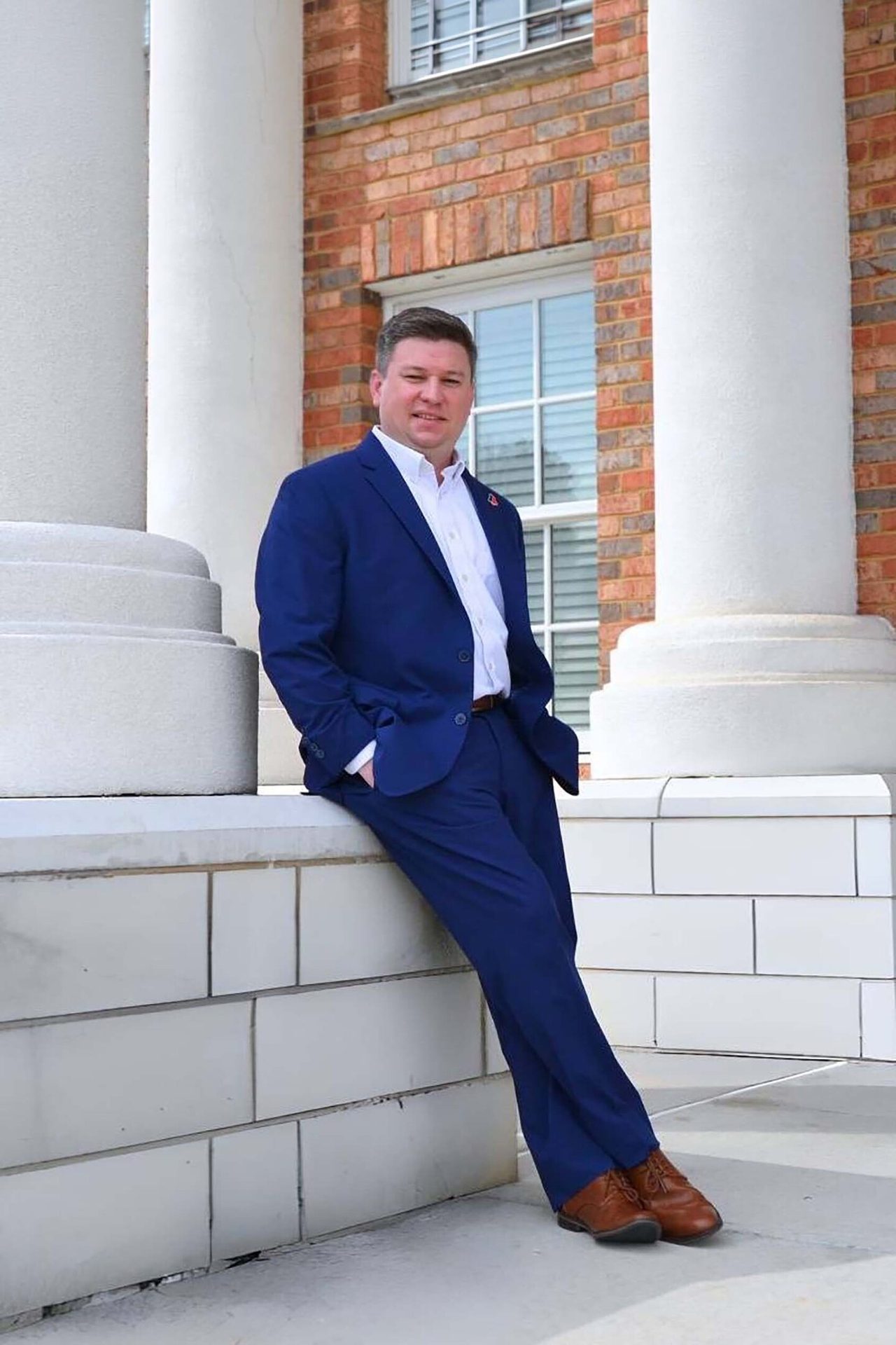 Man in blue suit leaning against a white wall with columns and a brick building in the background.