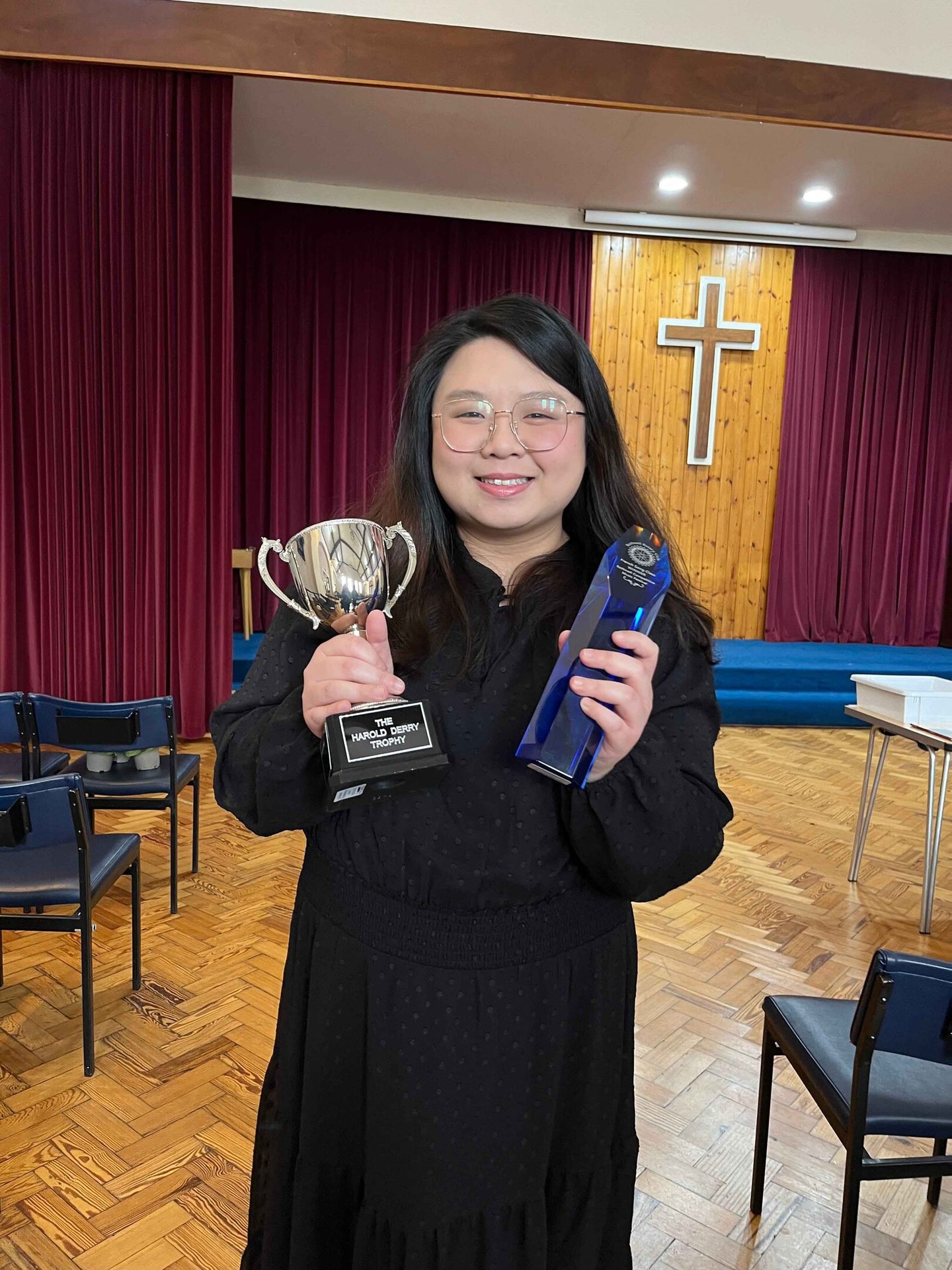 Smiling woman holding a silver trophy and blue award in front of a cross and red curtains.