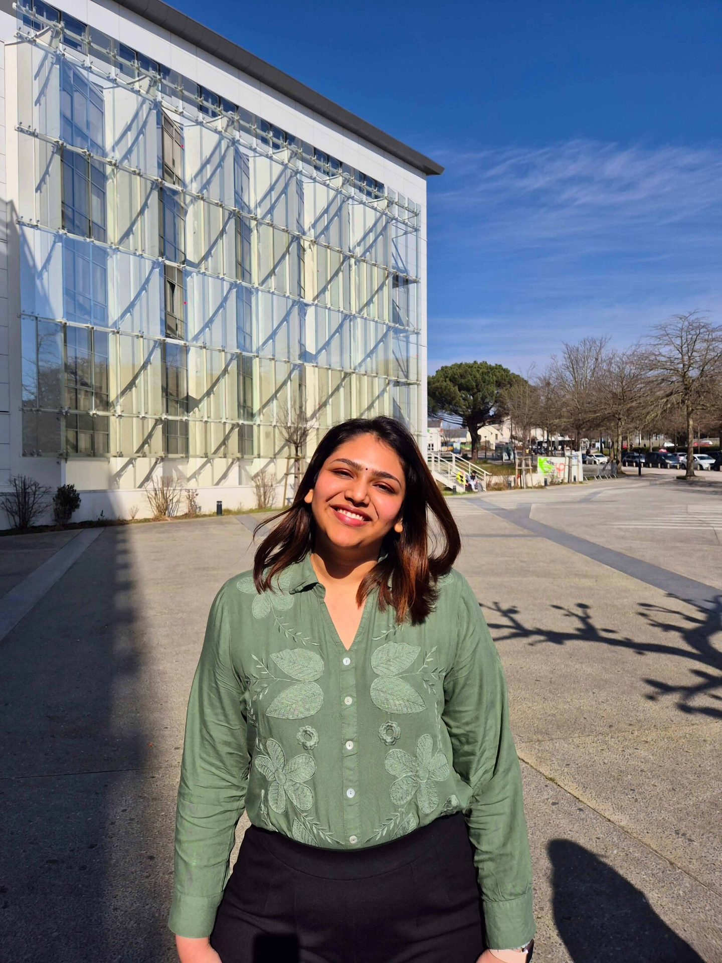 Smiling woman in a green embroidered shirt outdoors, in front of a modern glass building on a sunny day.