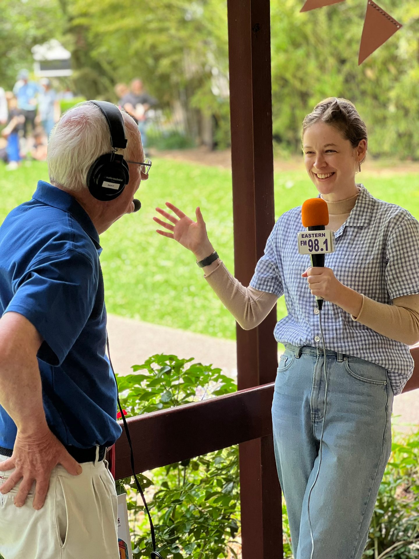 A man with headphones chats with a smiling woman holding an "Eastern FM 98.1" microphone outdoors.