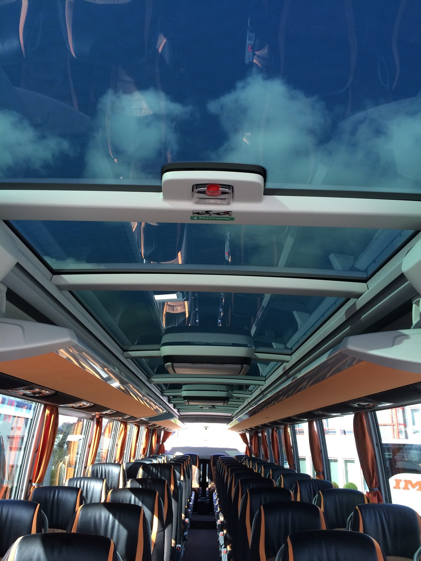 Interior of a modern bus with a panoramic glass roof showing the sky and rows of black and orange seats.