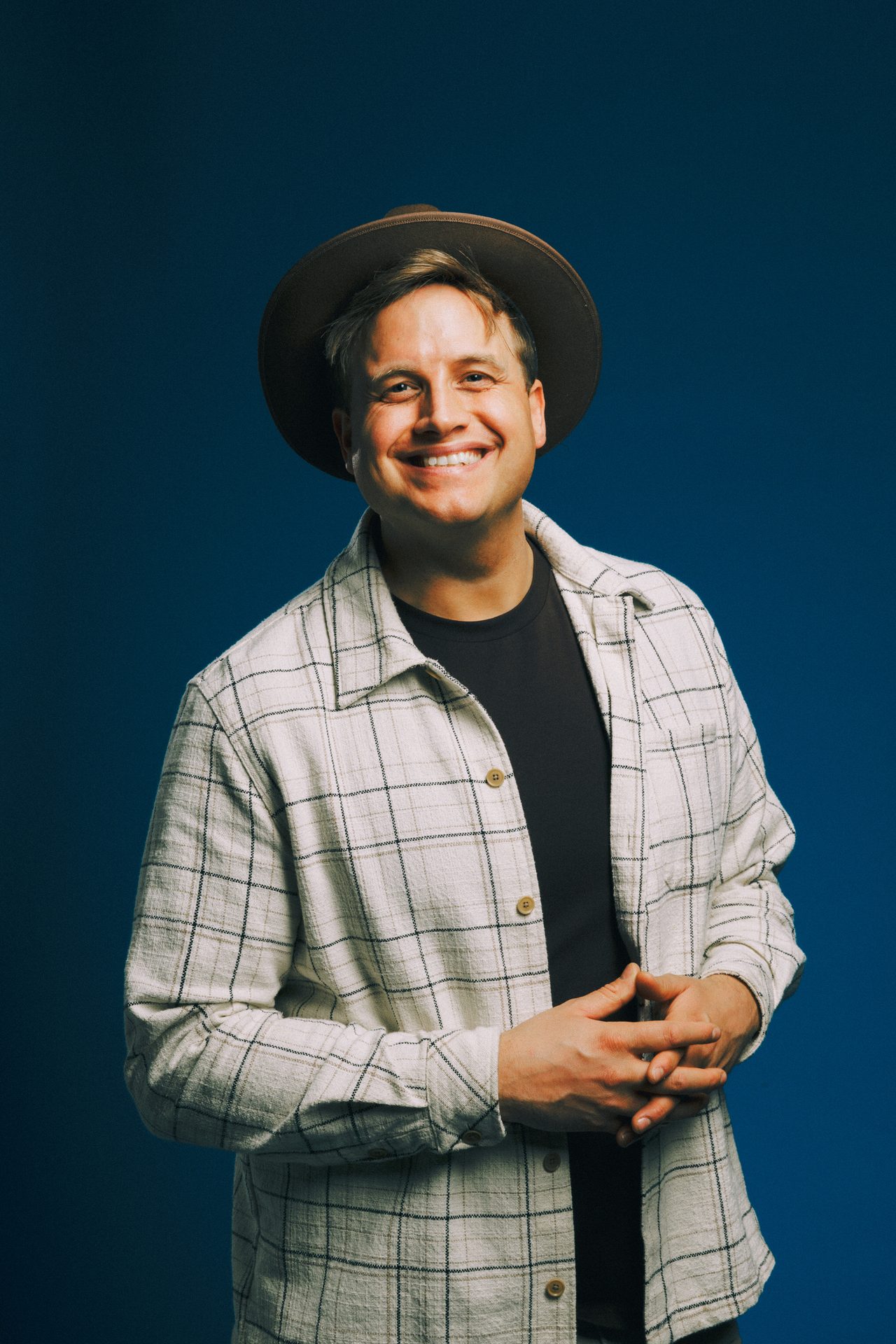 Smiling man in a plaid shirt and hat against a blue background.