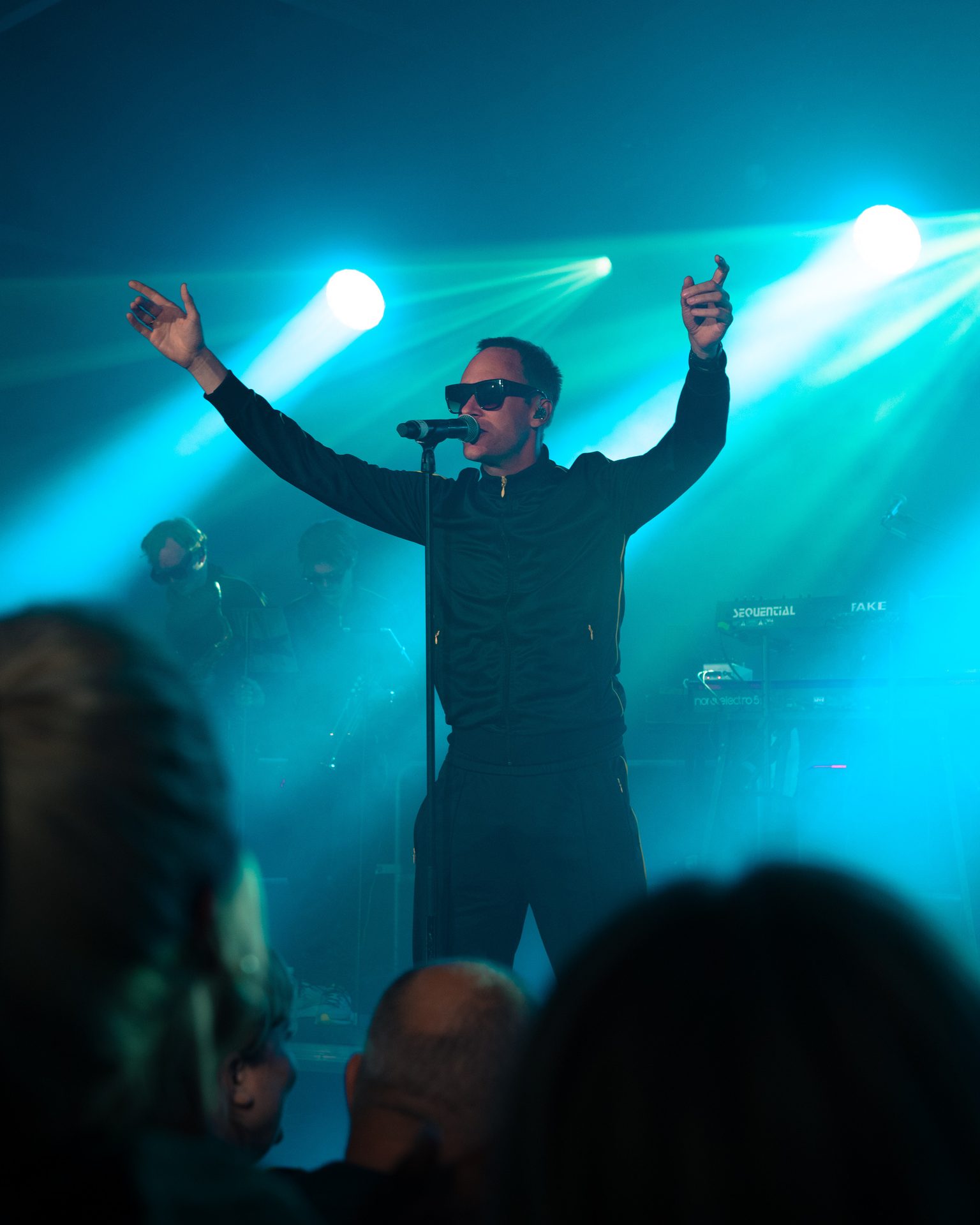 Singer in a black tracksuit and sunglasses, arms raised on stage under dramatic blue lights.