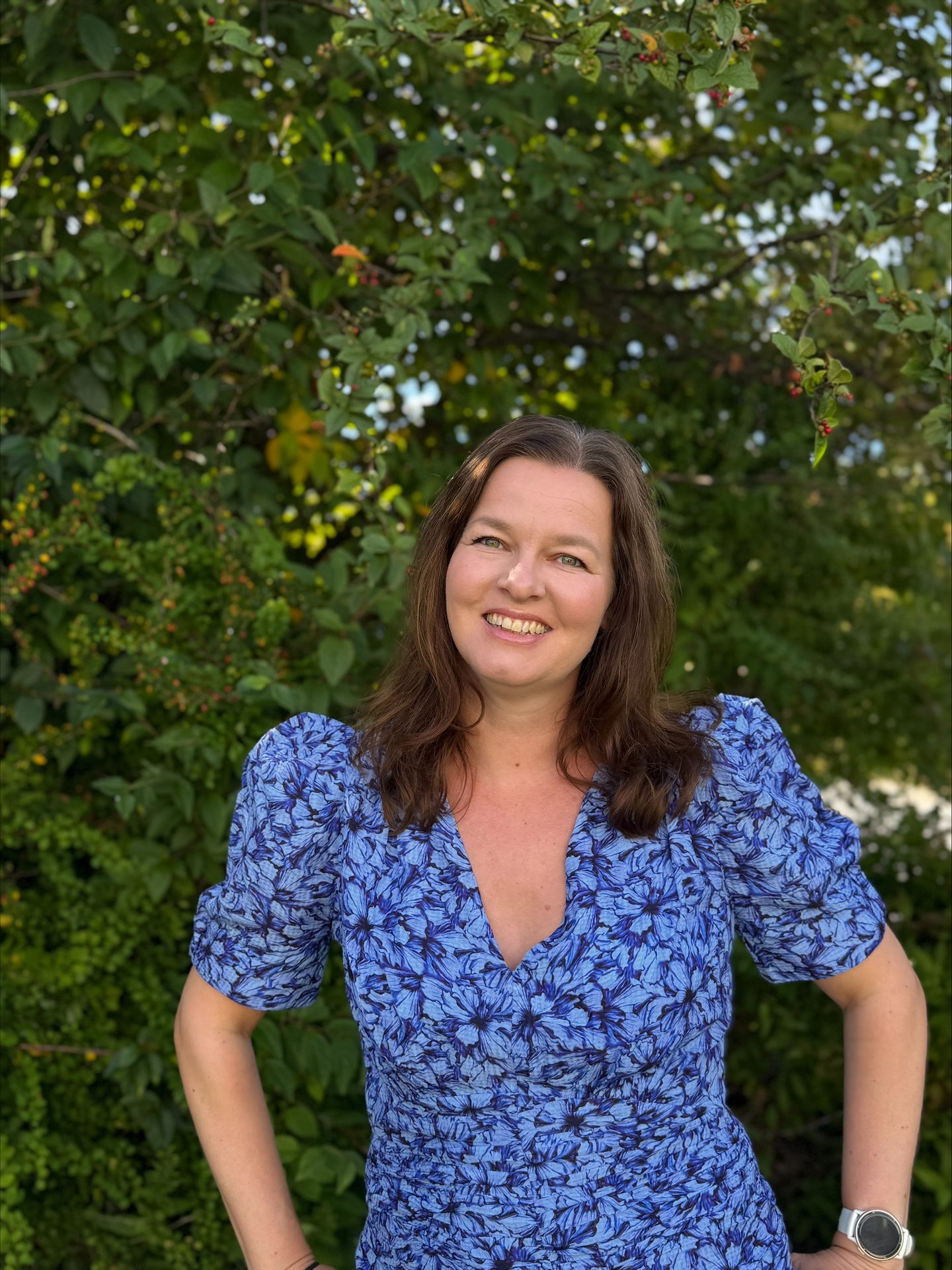 Smiling woman in a blue floral dress posing against a green leafy background on a sunny day.