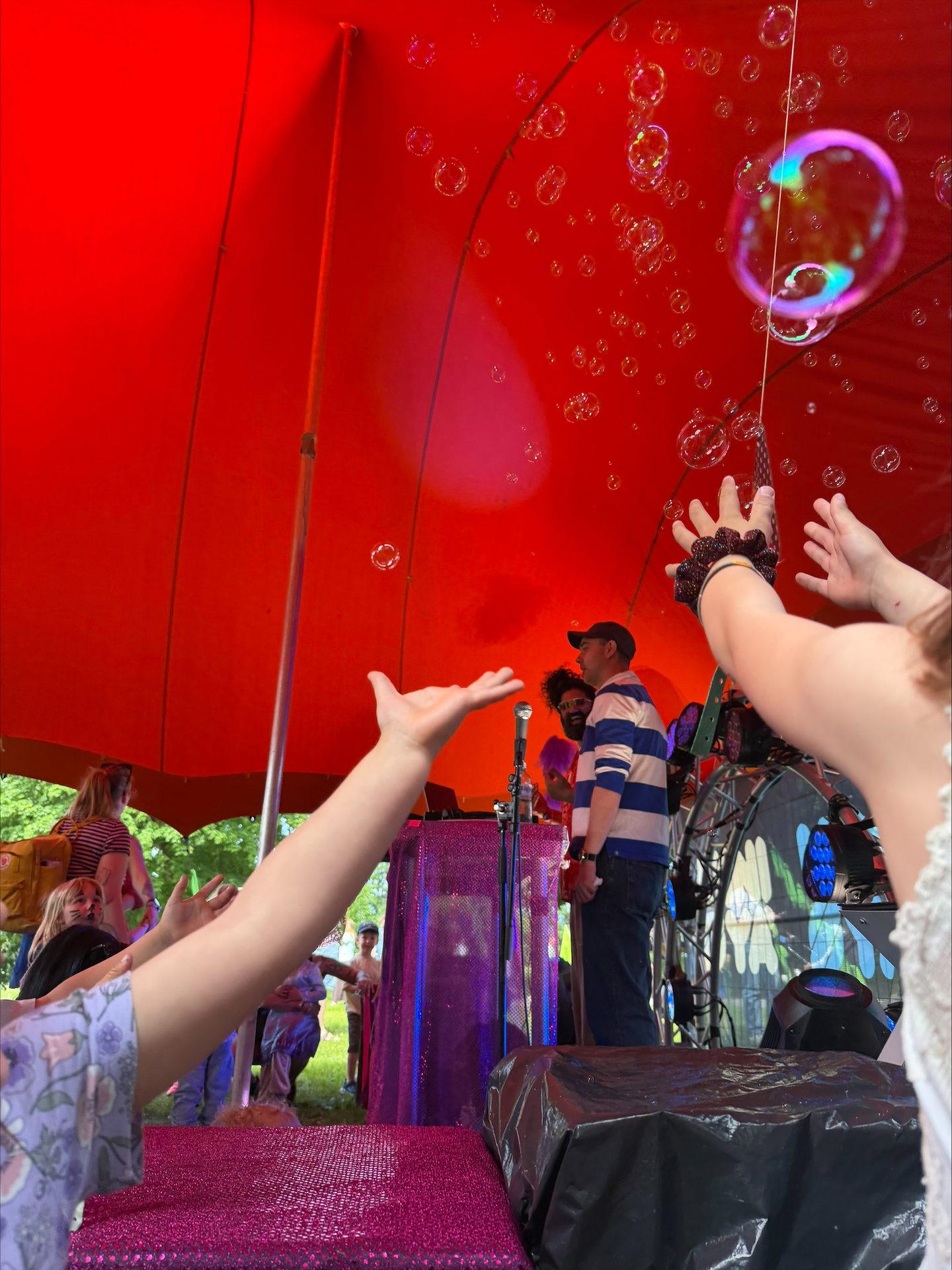 Children's hands reach for bubbles under a red tent with performers on a glittery stage.