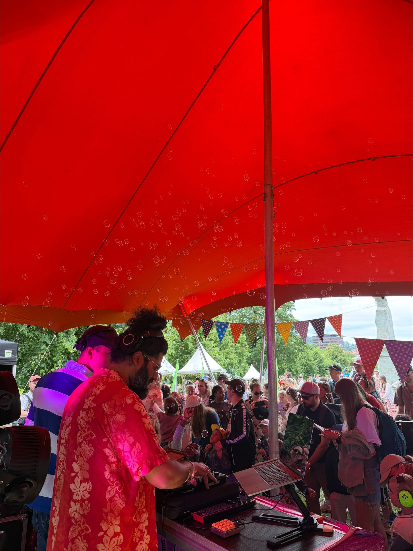 DJ in red floral shirt playing music under a red canopy with headphones, surrounded by bubbles and a crowd.