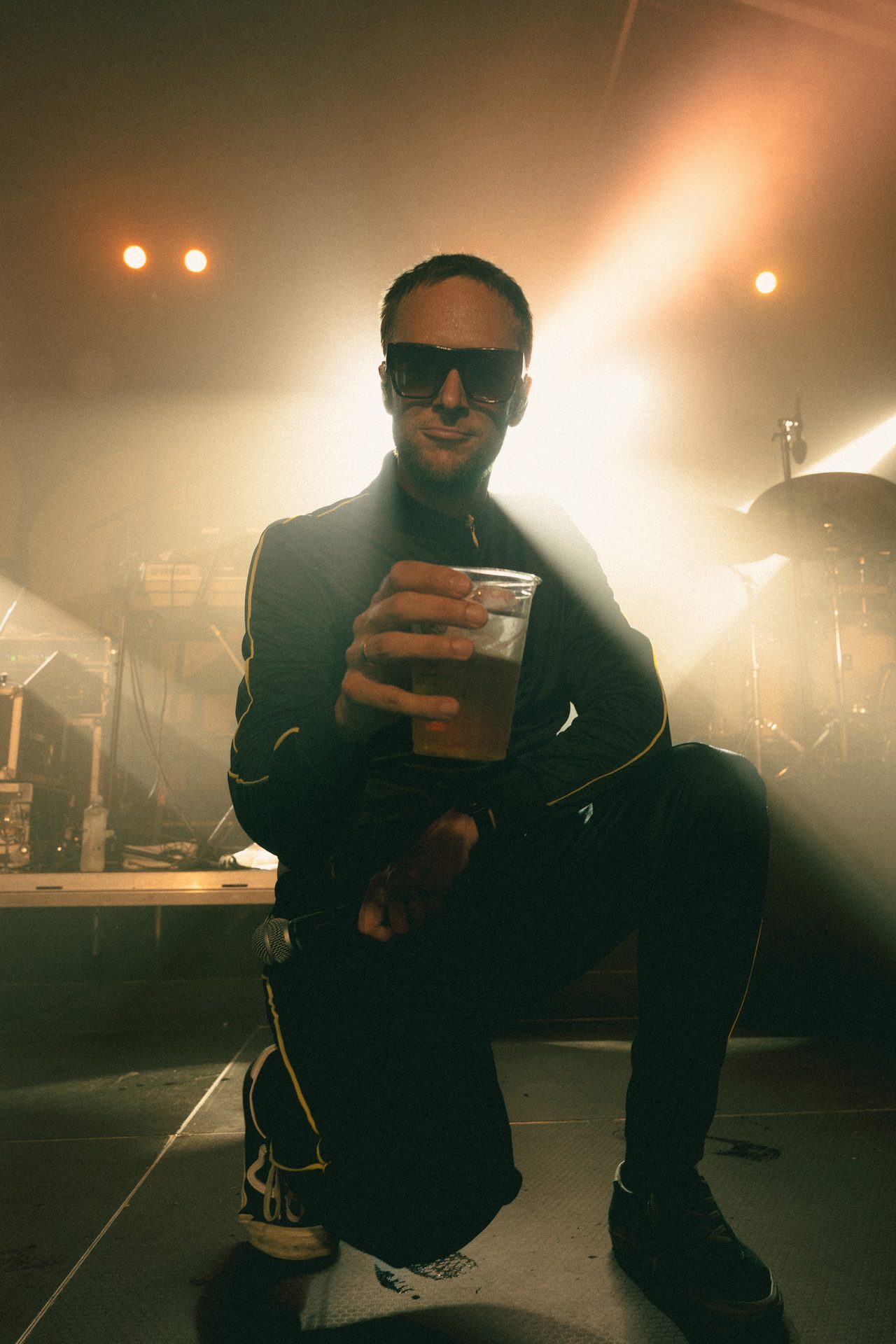 A man in sunglasses and a tracksuit kneels on a stage, holding a drink, under bright lights.