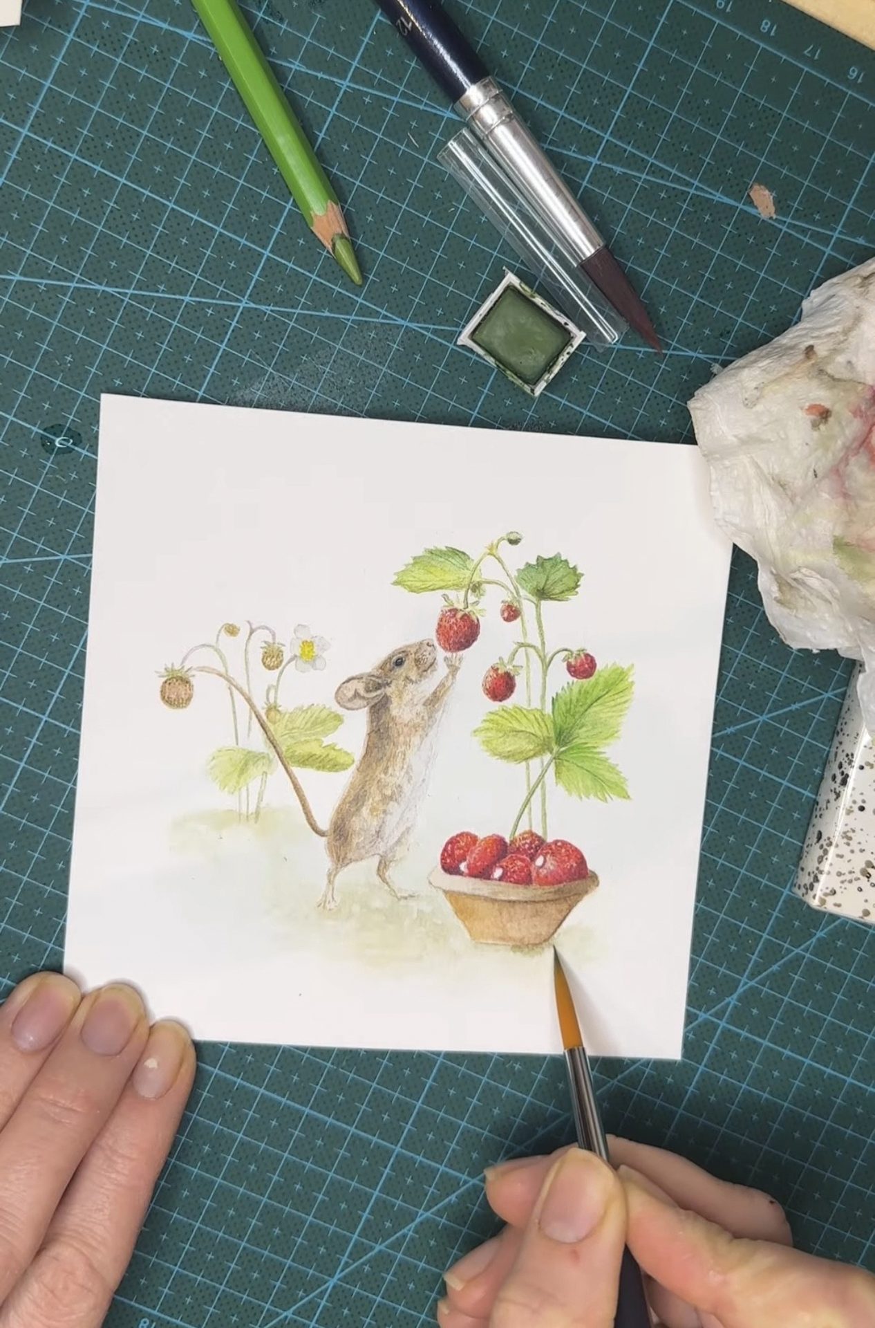 Close-up of hands painting a watercolor illustration of a mouse reaching for strawberries on a white paper.