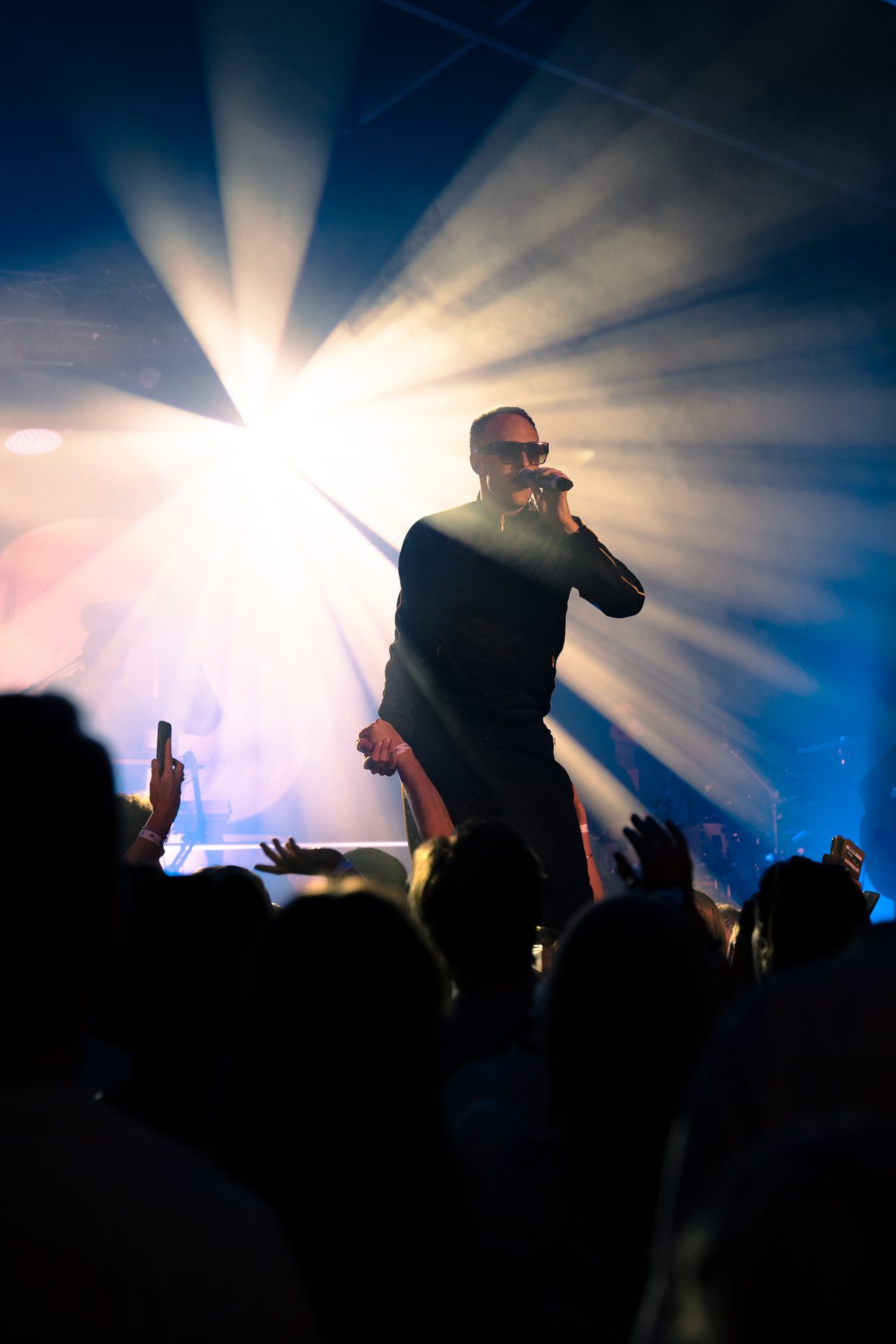 A performer in sunglasses sings on a brightly lit stage, interacting with the silhouetted audience.