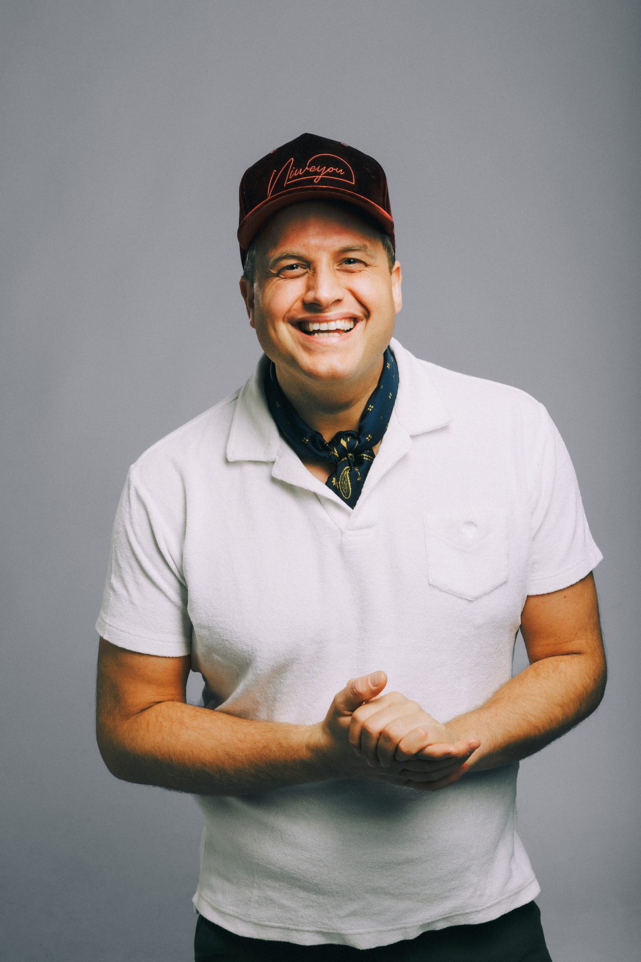 Smiling man in cap, white polo, neckerchief, hands clasped, against a gray background.