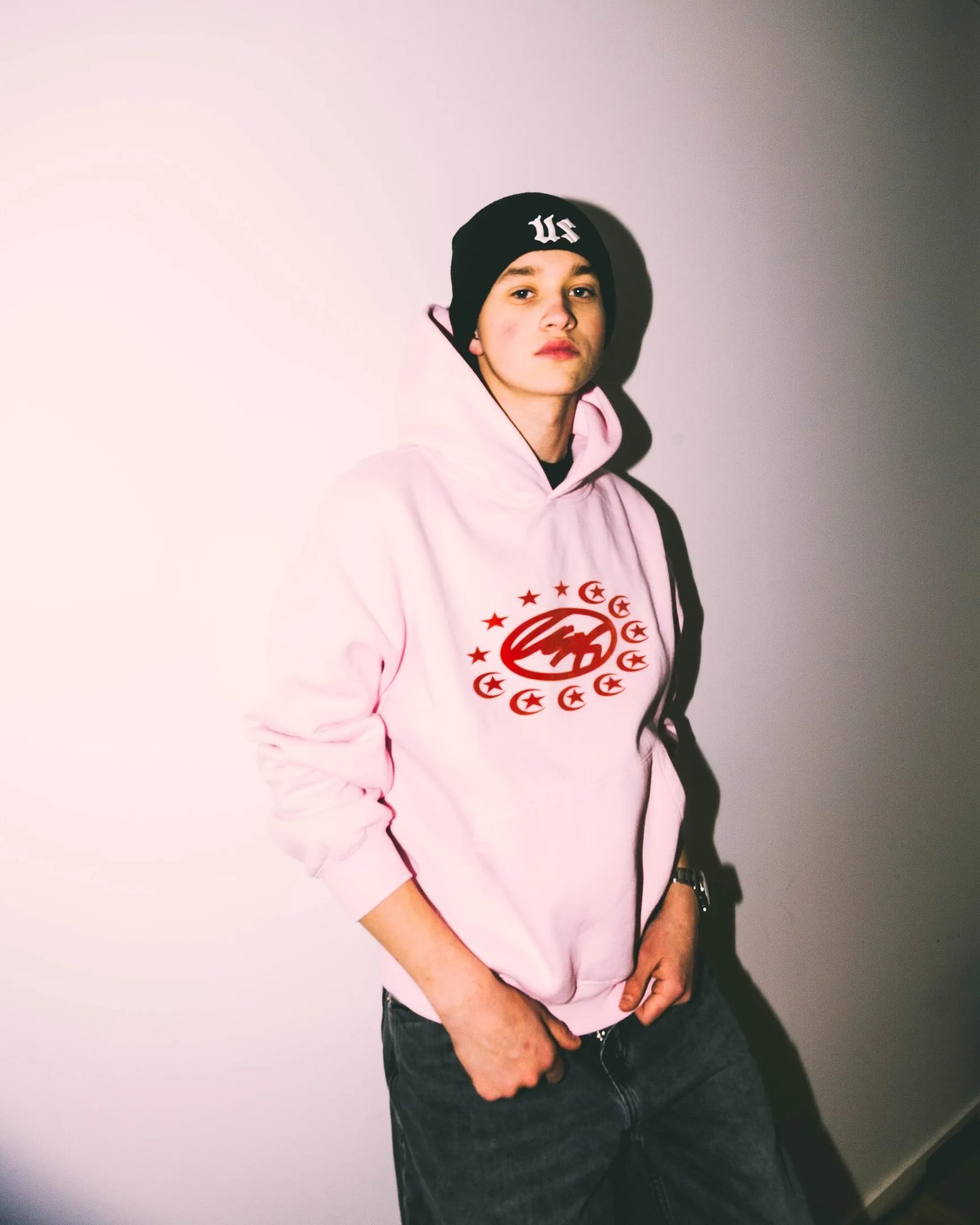 Young person wearing a pink hoodie with a red logo, black beanie, and jeans, posing against a white wall.