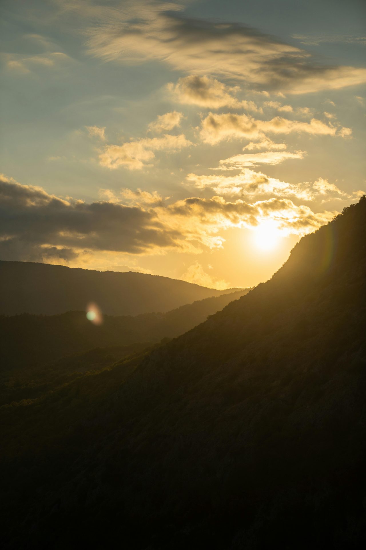 Golden sunset behind dark mountain silhouettes, with bright sun and clouds.