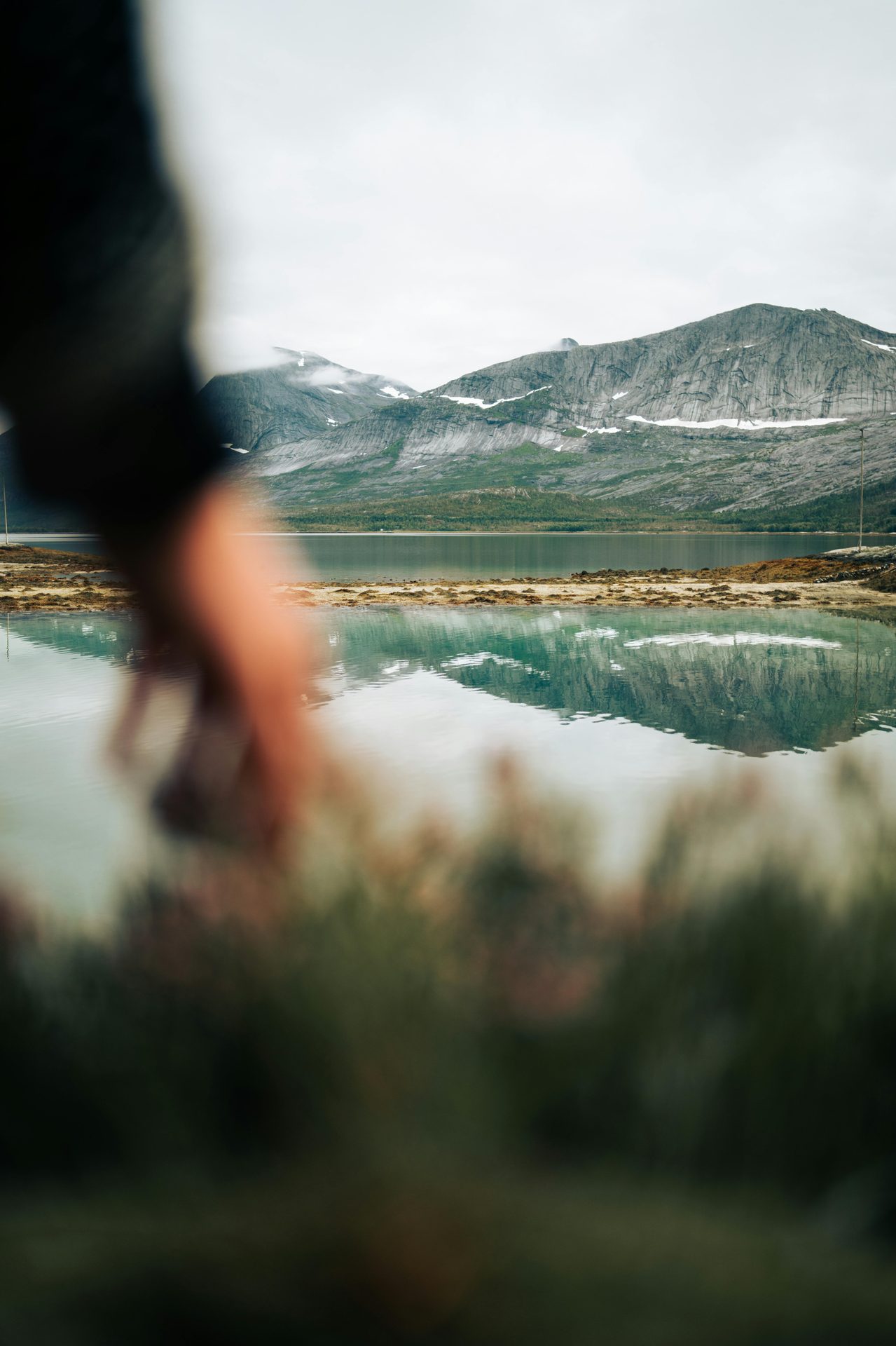 Blurred arm reaching, lake reflects mountains under cloudy sky.