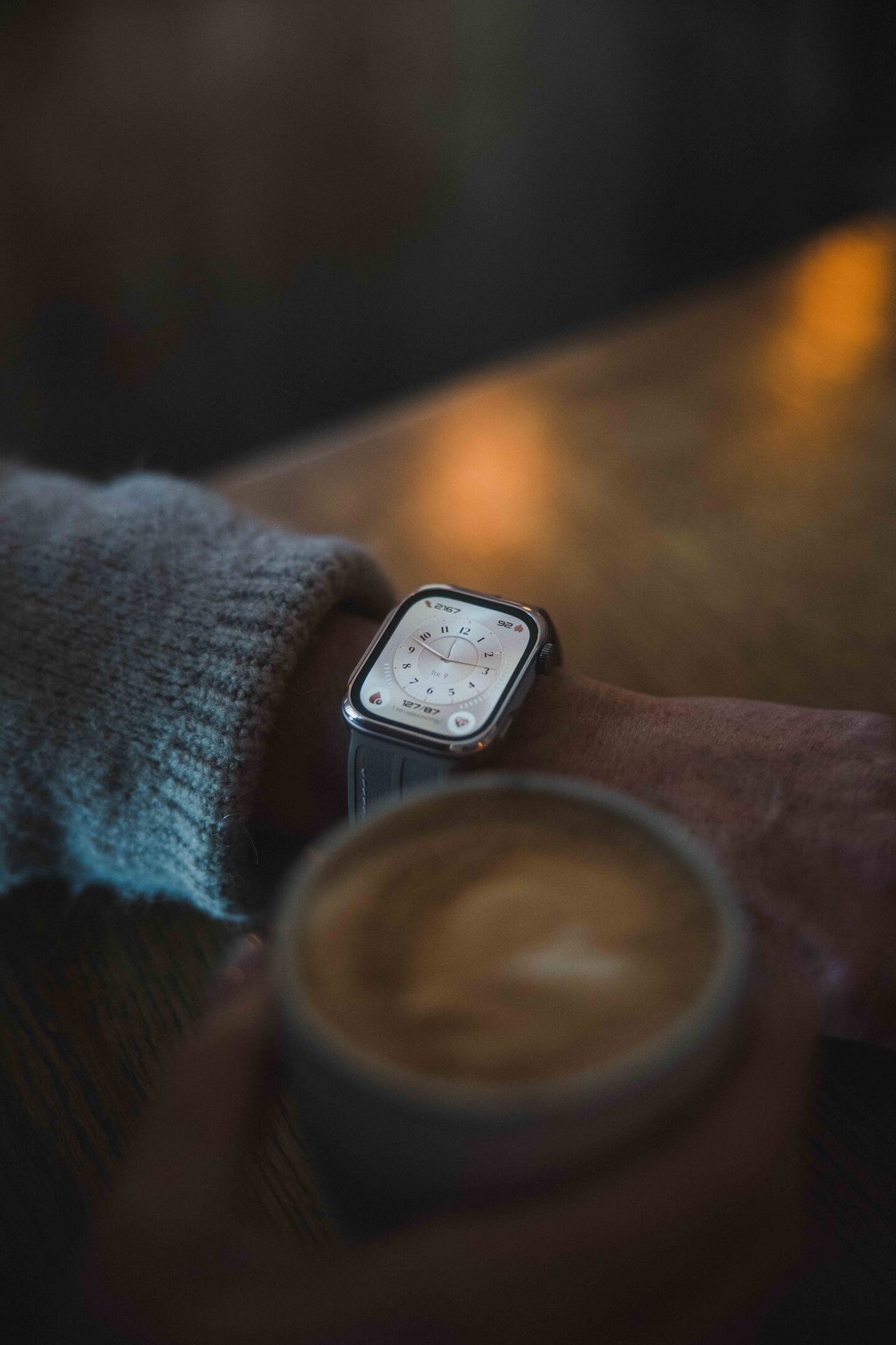 A person wearing an Apple Watch holds a coffee cup, showing the watch face and a latte.