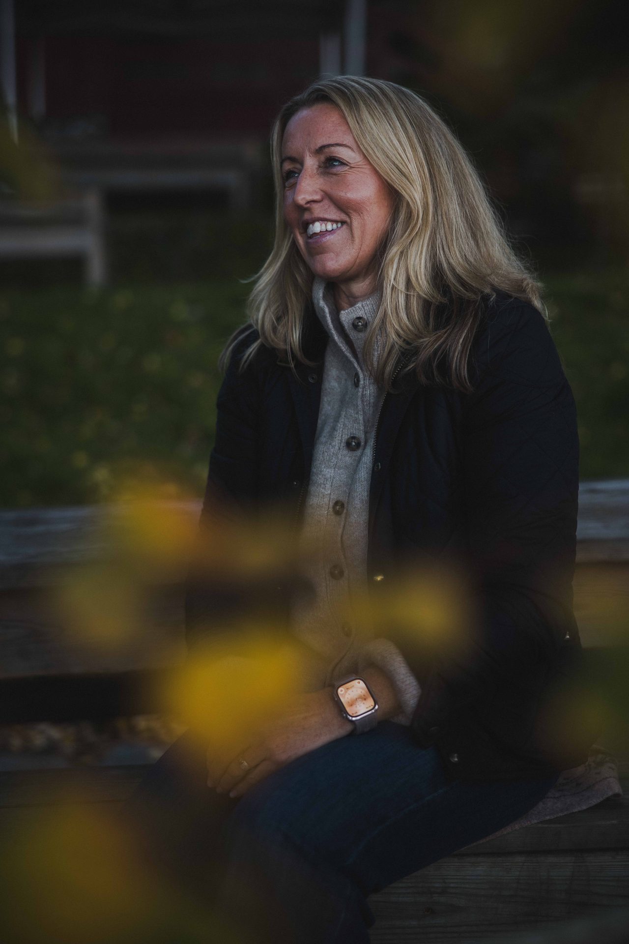 A blonde woman in a dark jacket and grey sweater smiles while looking to the side, with blurred yellow leaves in the foreground.