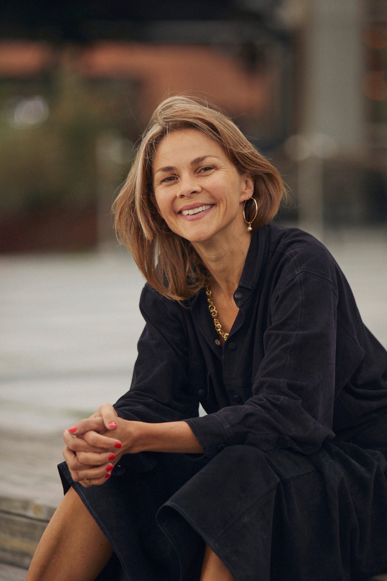 Happy woman with light hair, gold jewelry, and black attire, sitting outdoors.