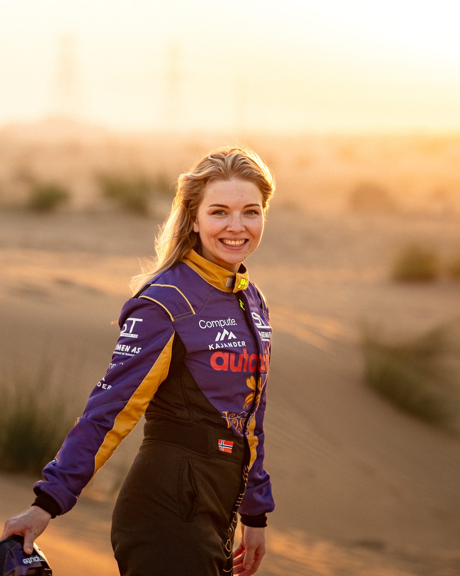 Smiling blonde woman in a purple and yellow racing suit, holding a helmet, in a desert at sunset.