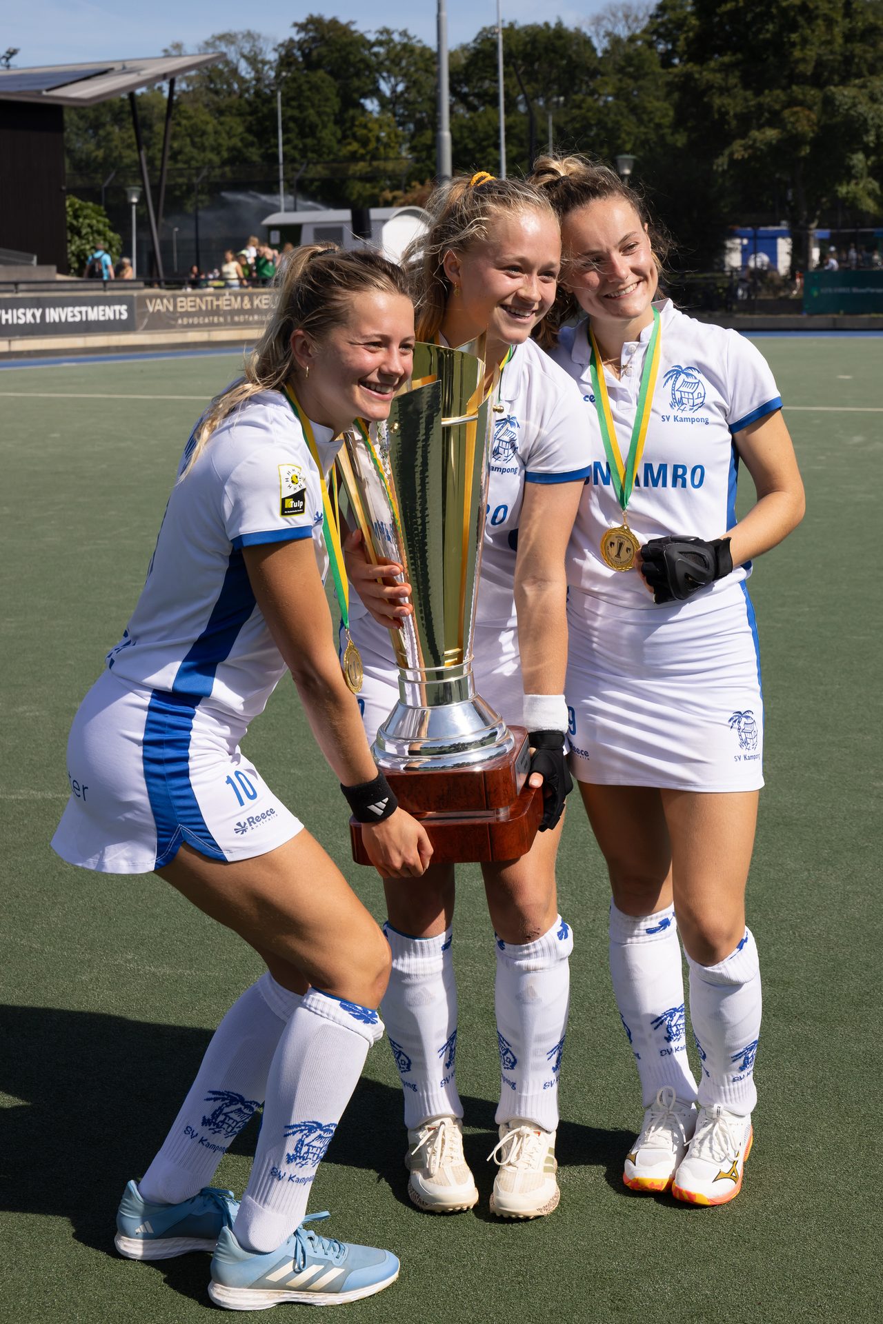 Three smiling female hockey players in uniform celebrating with a large gold trophy on a field.