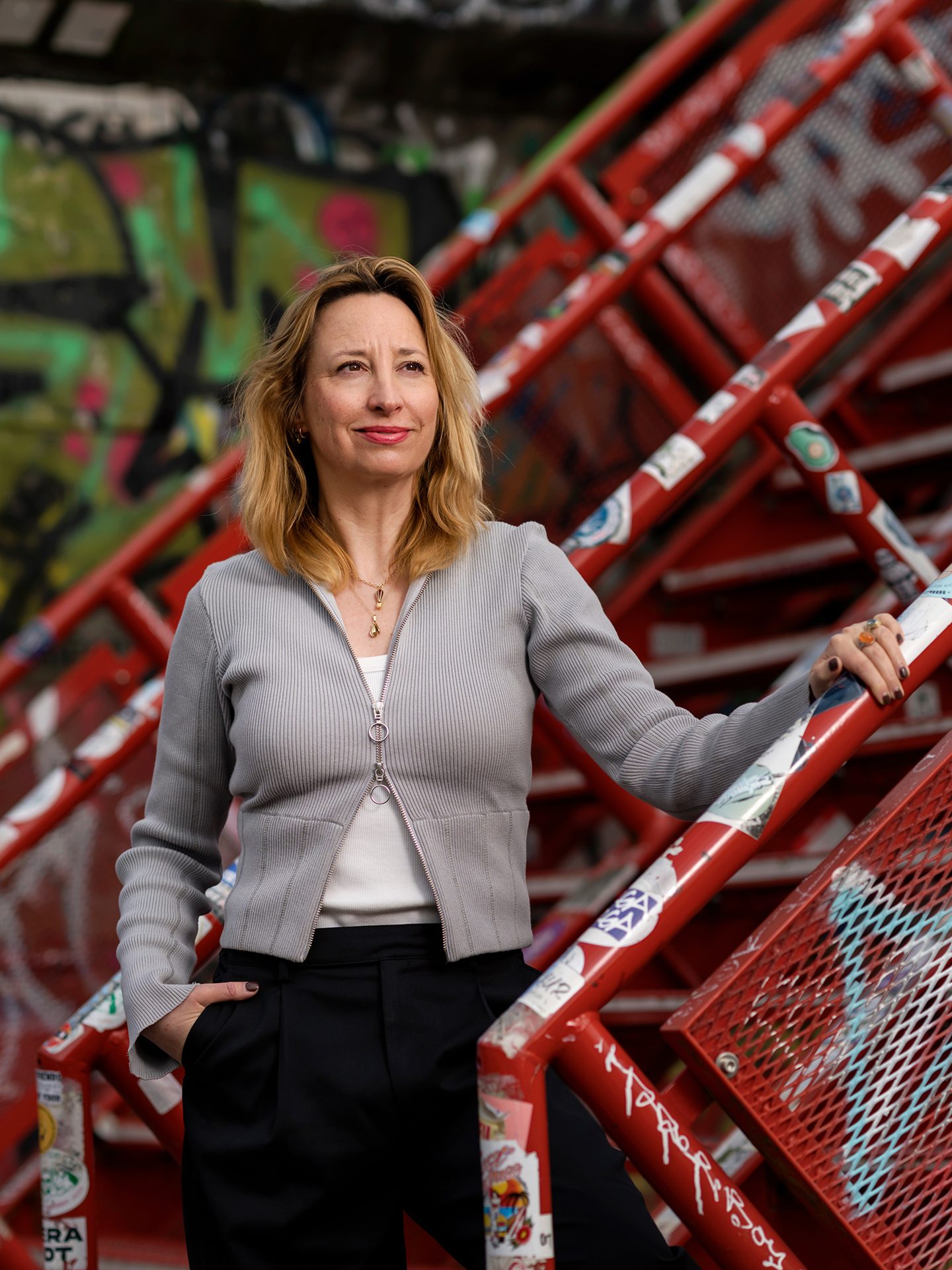 Woman in grey cardigan and black pants next to red railings with graffiti, looking up.