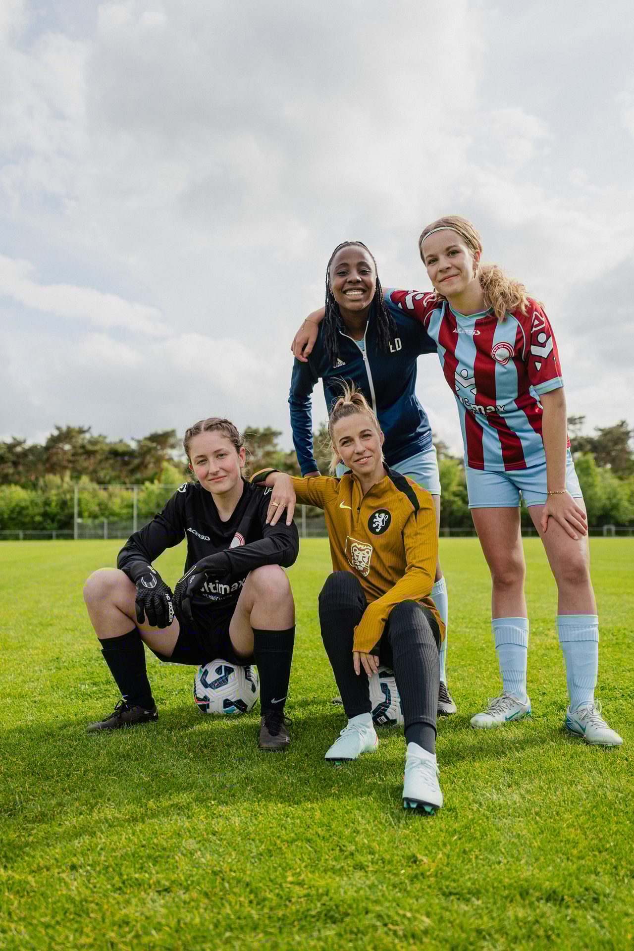 Four smiling female soccer players posing on a field, two standing, two crouching.