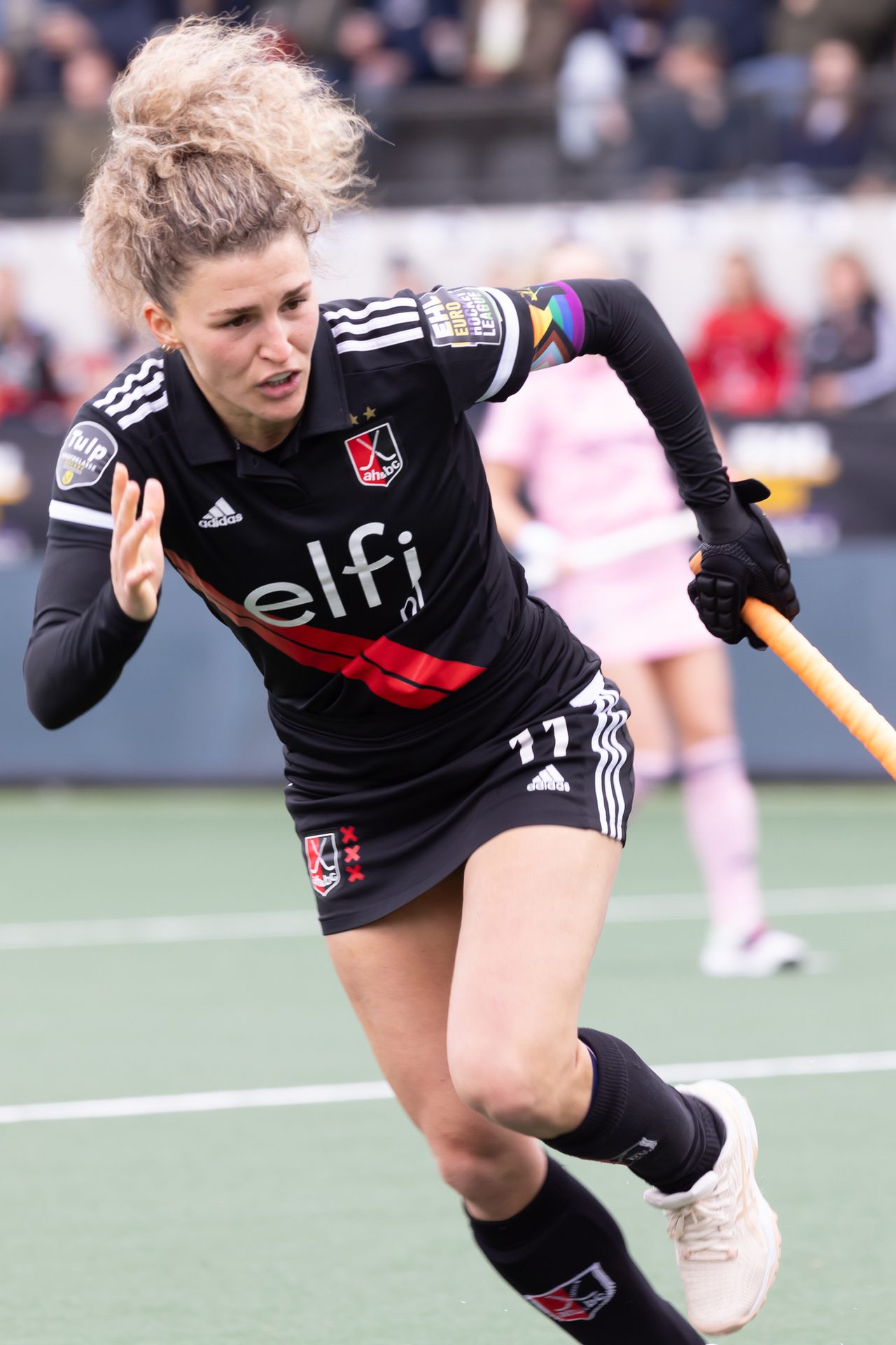 A female field hockey player with curly hair in a black uniform runs with a stick on a green field.
