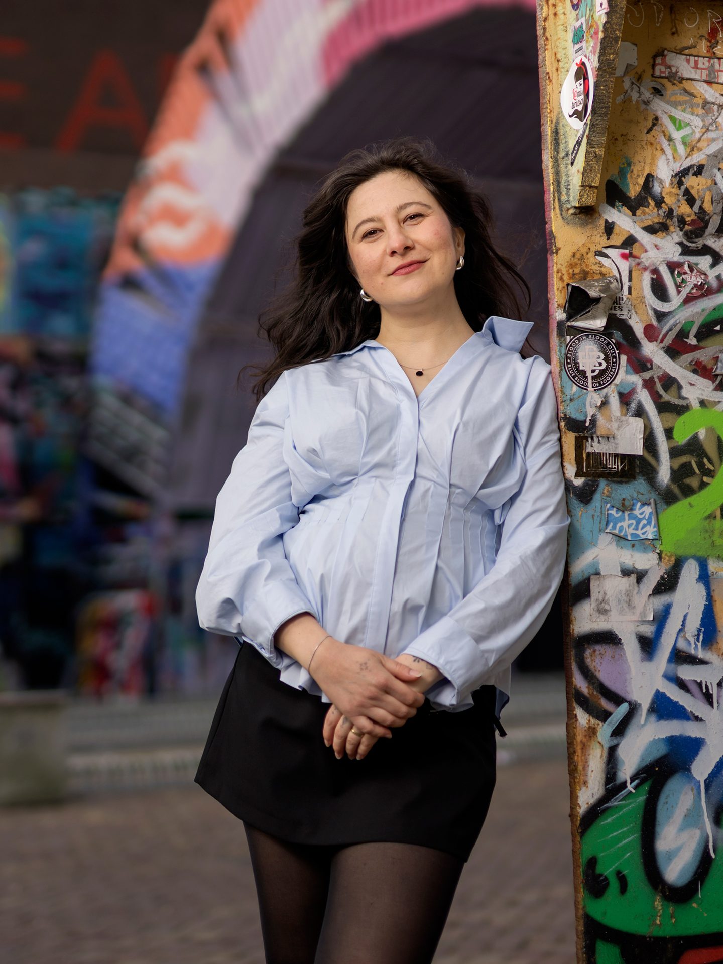 Woman in blue shirt and black skirt leans on a graffiti post, smiling, with a colorful blurred background.