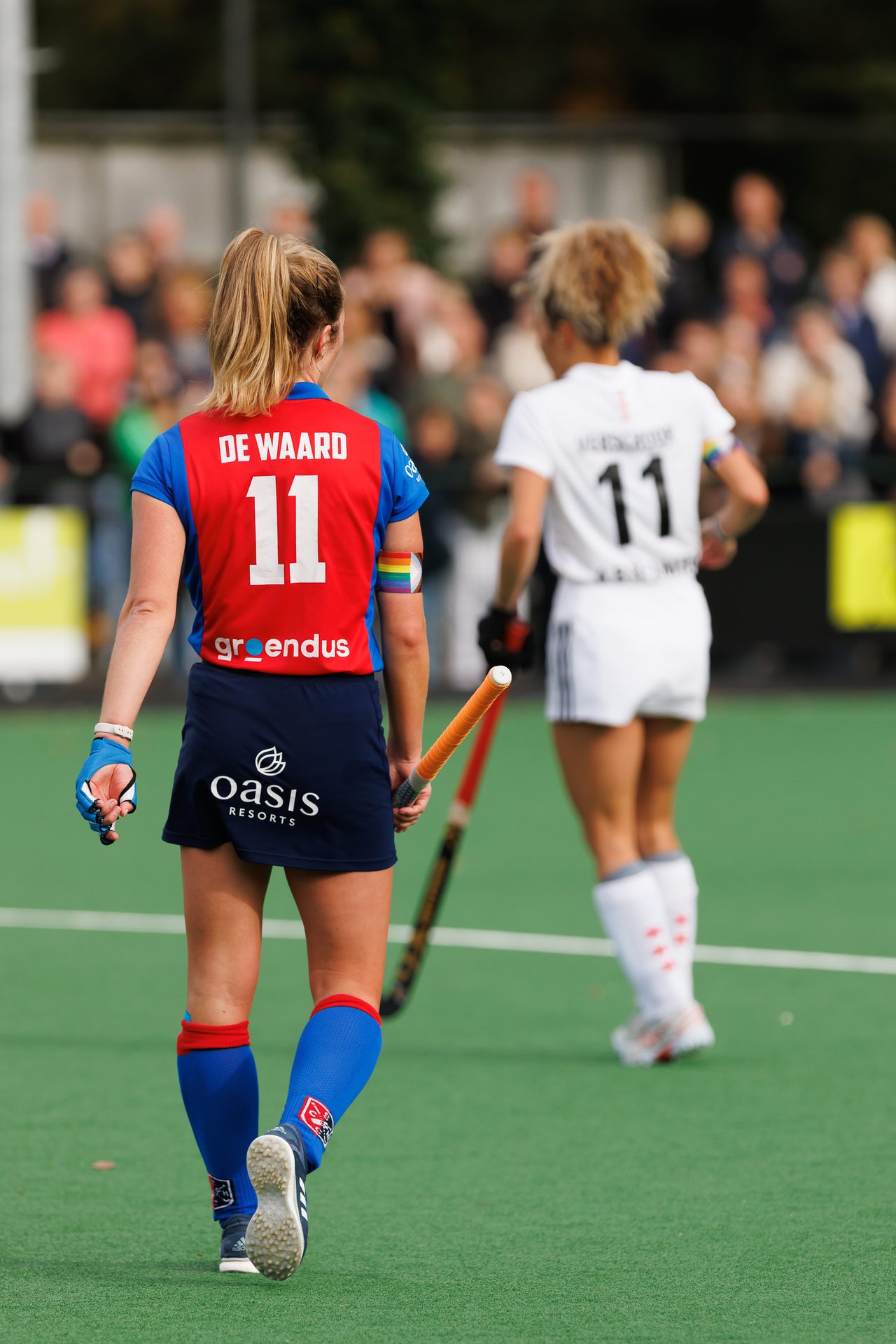 Two female field hockey players, one in red/blue "DE WAARD 11" with a rainbow armband, on a green field.