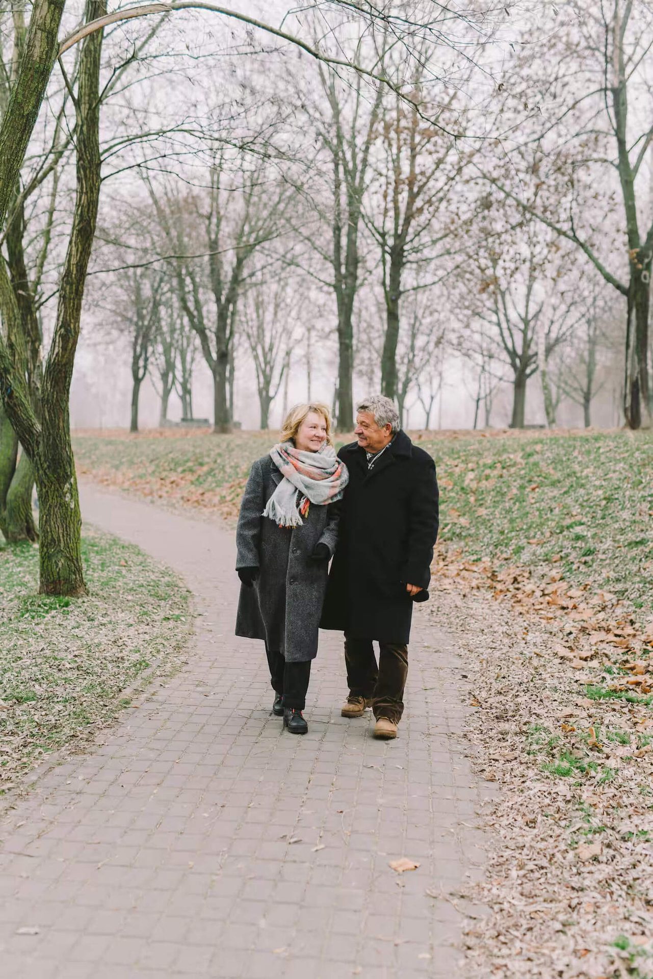 Happy elderly couple walks on a park path, smiling at each other among bare trees.