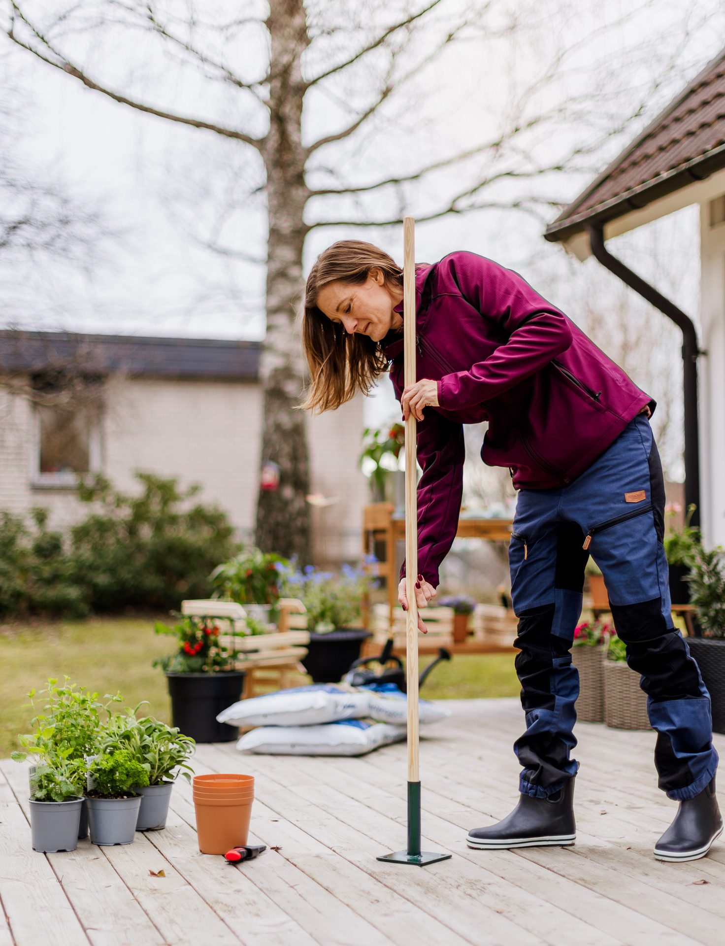 A woman in a purple jacket and work pants bends over a gardening tool on a wooden deck, surrounded by potted plants and bags of soil, preparing for spring planting.