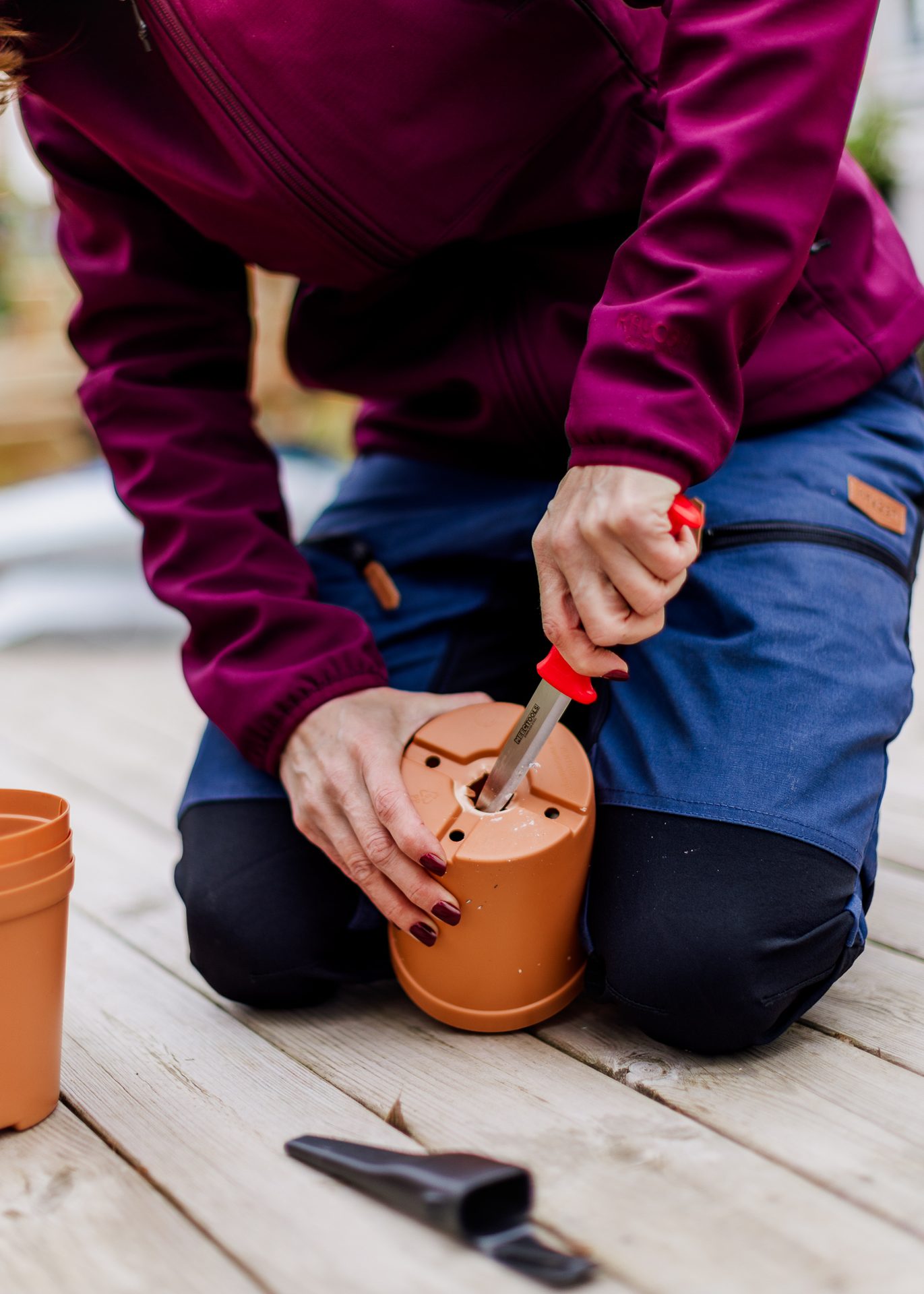 Person kneeling, using a red-handled knife to make a hole in the bottom of a terracotta-colored plant pot.