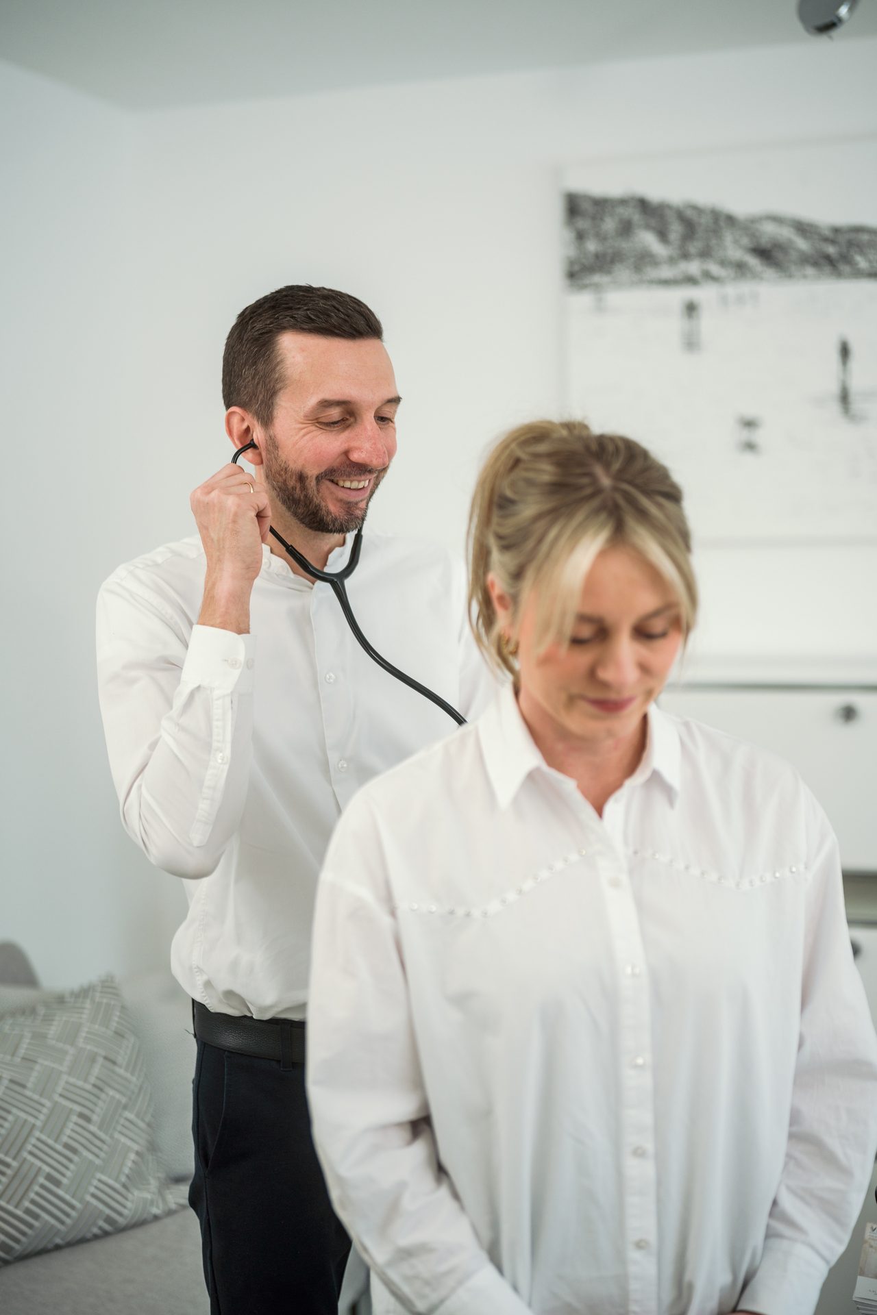 A smiling male doctor examines a female patient's back with a stethoscope during a medical check-up.