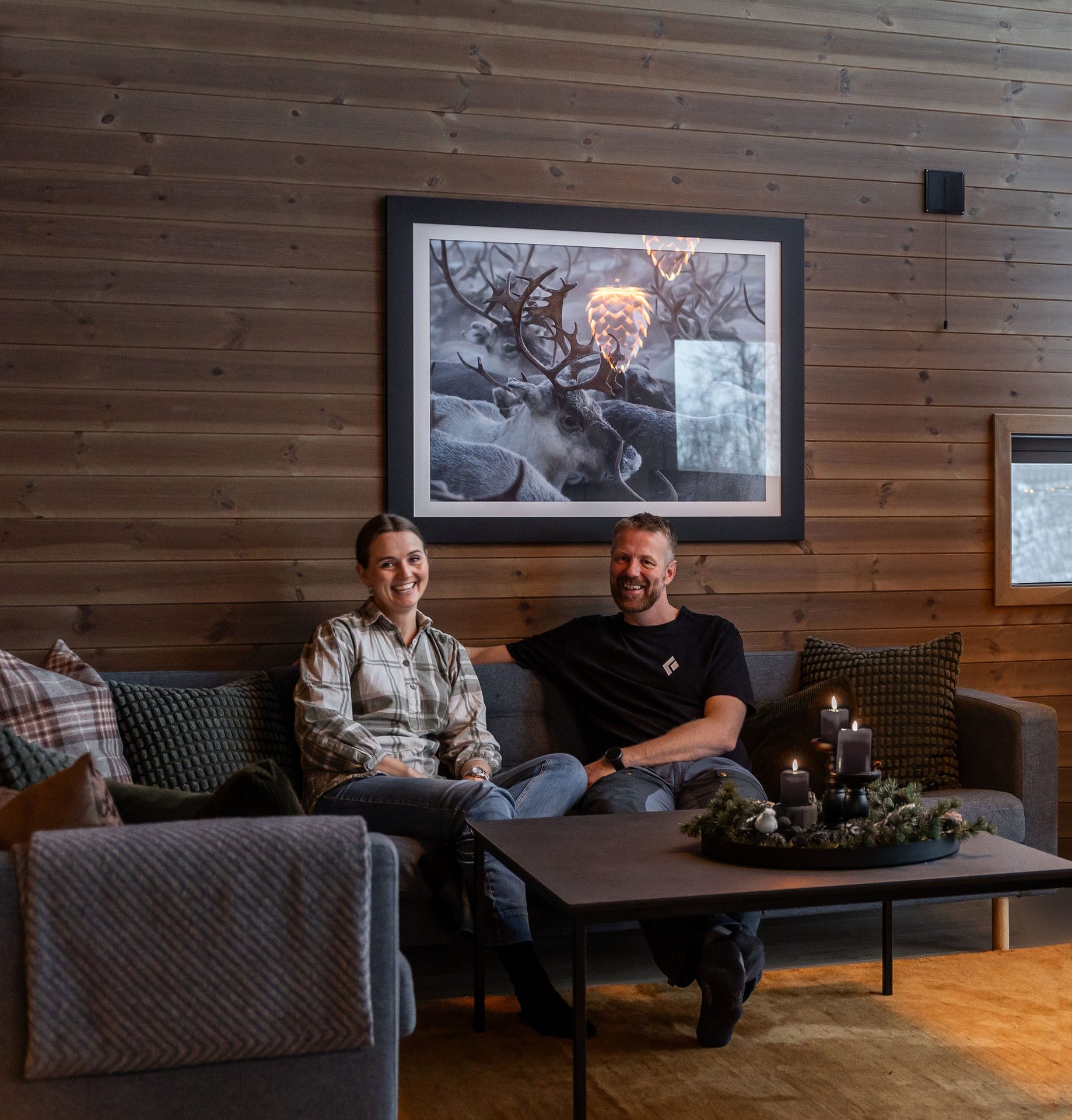 Two happy people on a couch in a cozy room with wood walls, a reindeer picture, and candles.