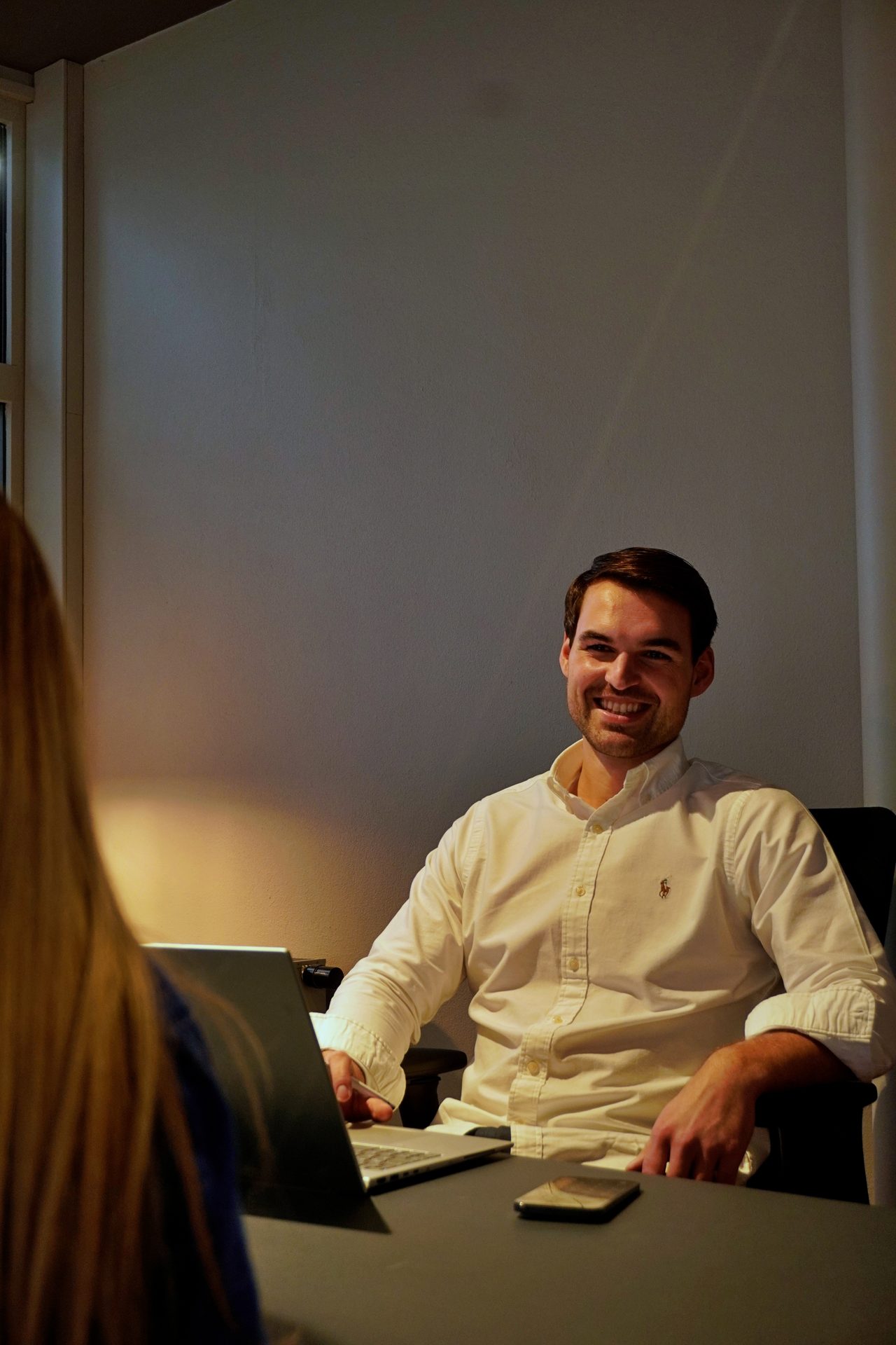 Smiling man in white shirt at desk with laptop, facing another person.
