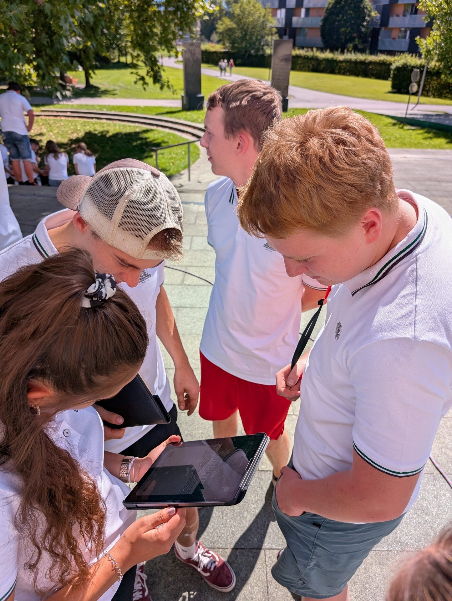 Four young people outdoors, gathered around a tablet on a sunny day.