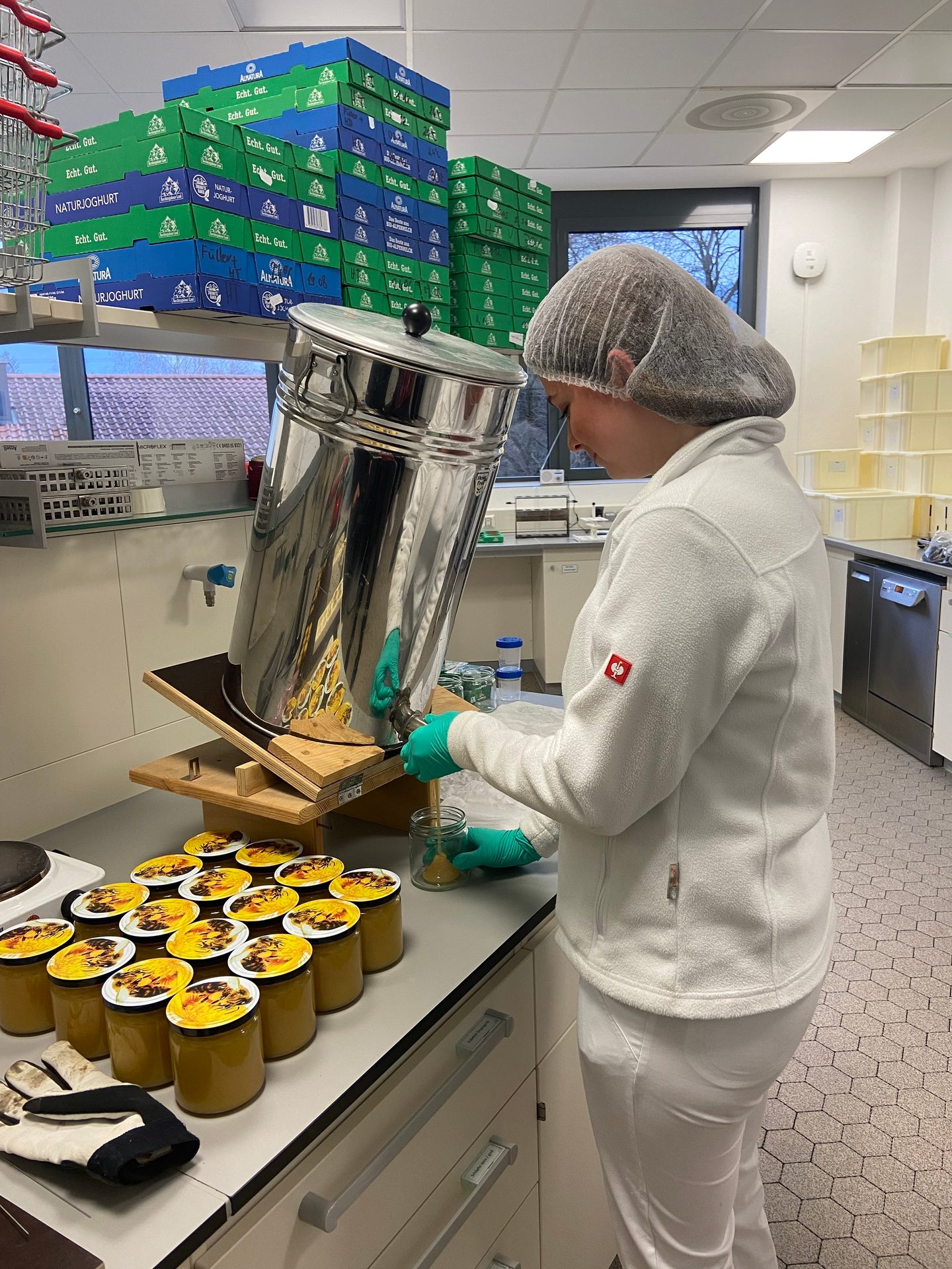 Person in lab gear filling jars with yogurt from a large dispenser in a production area.