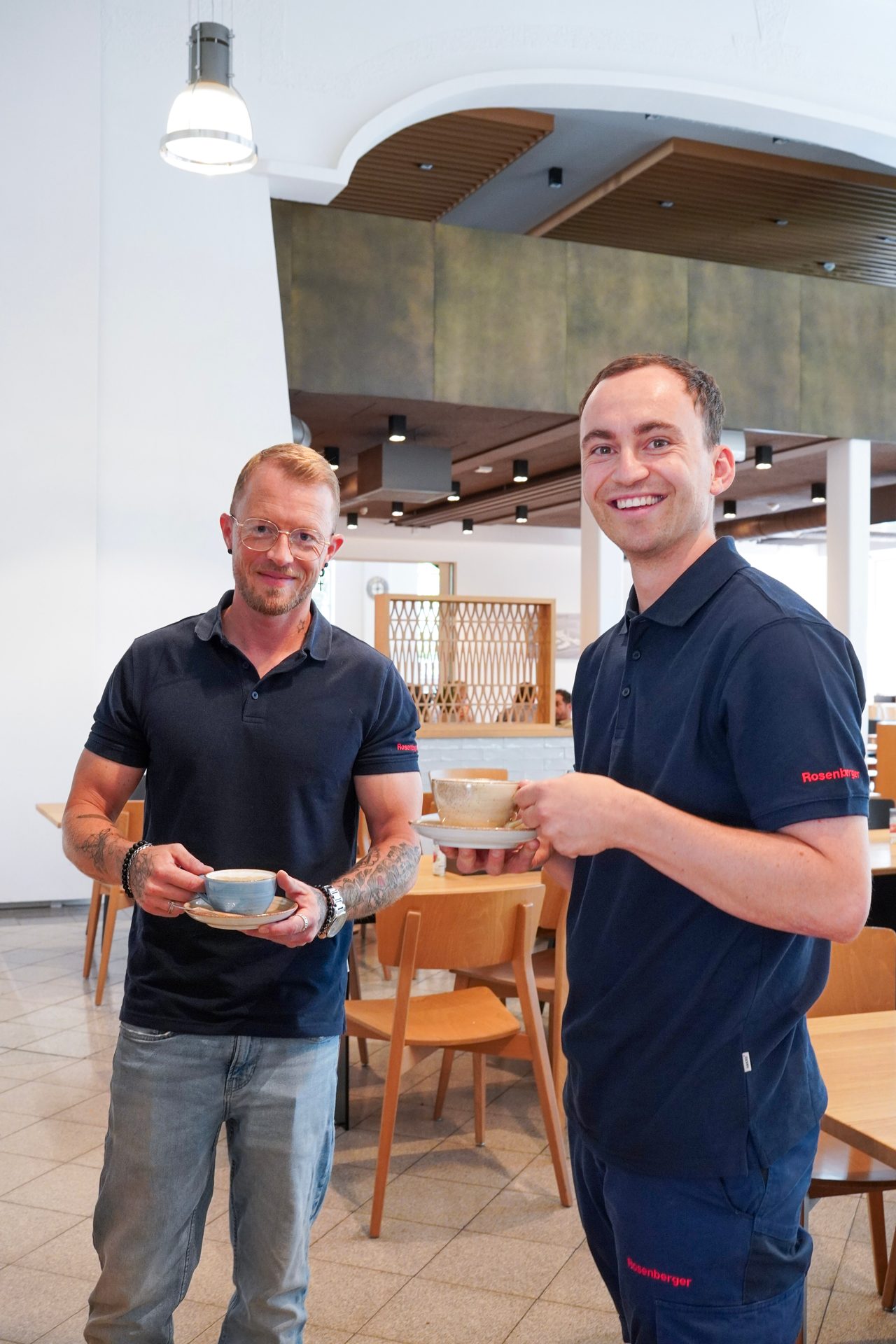 Two men in blue polo shirts hold coffee cups and smile in a modern cafe or office lounge.