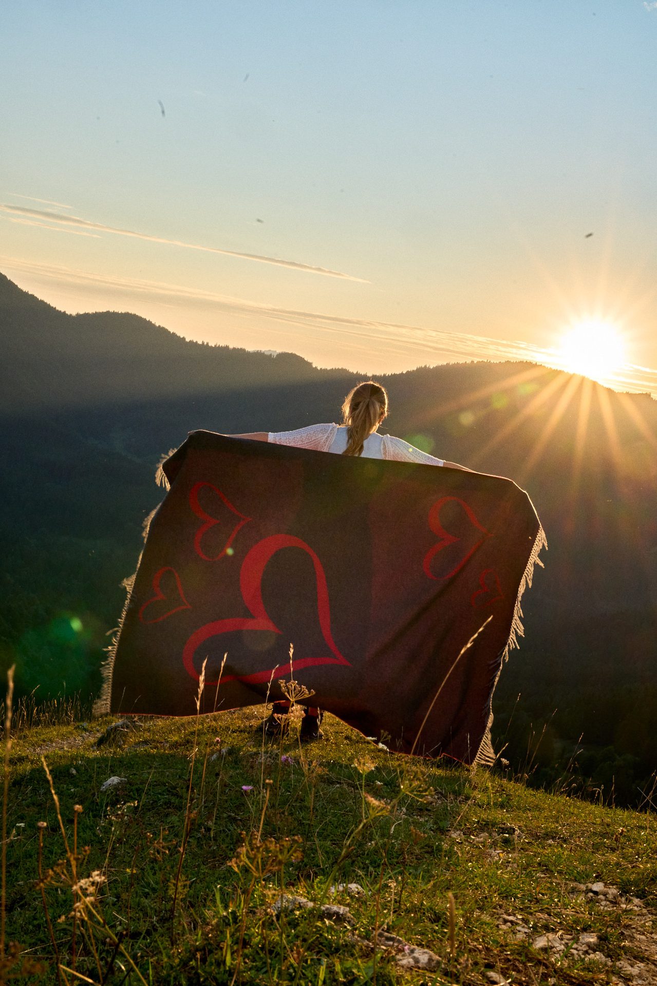 Woman on a hilltop, arms outstretched with a heart blanket, watching the sunset over mountains.