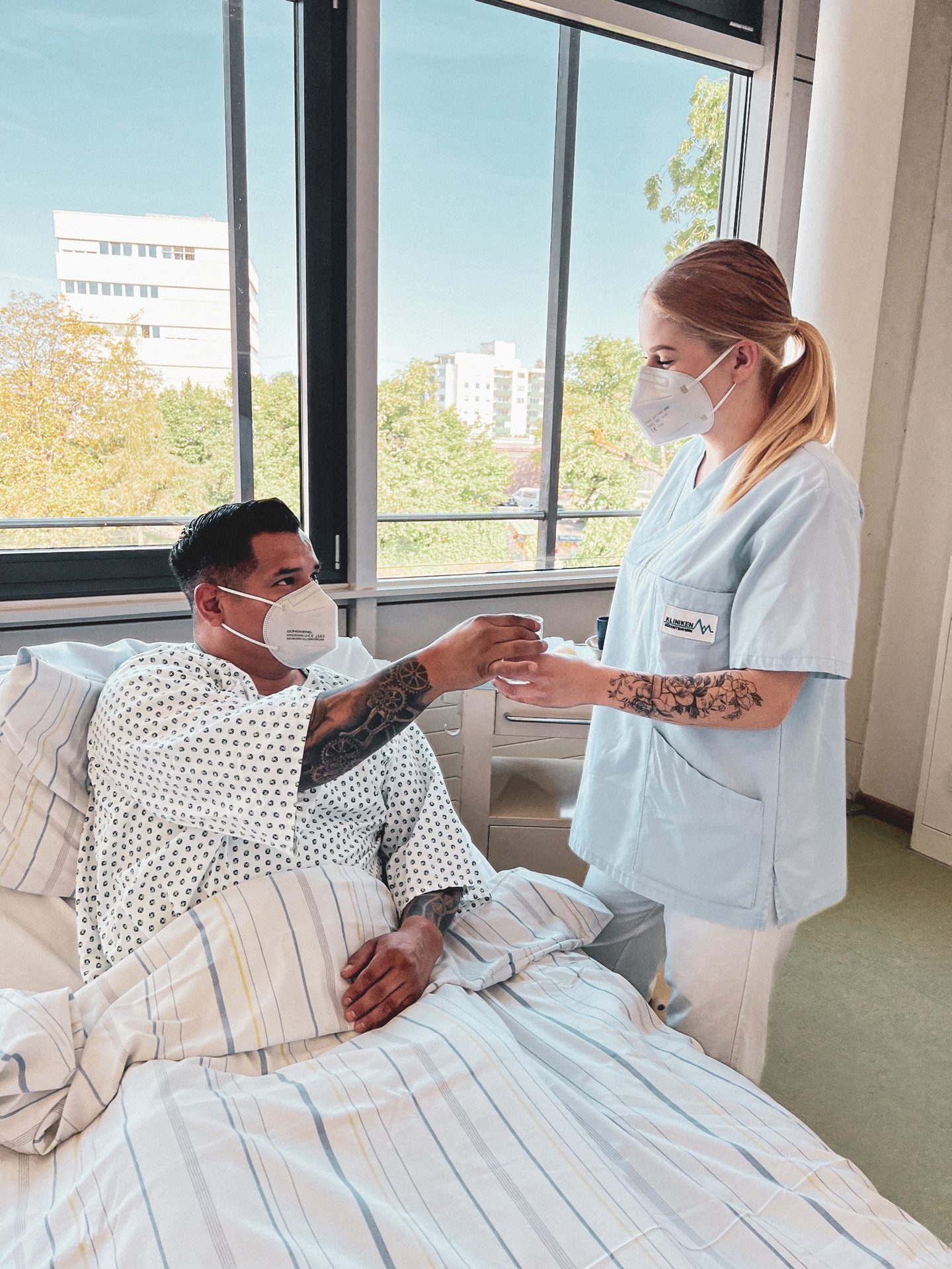 Masked nurse hands water to masked patient in a hospital bed.