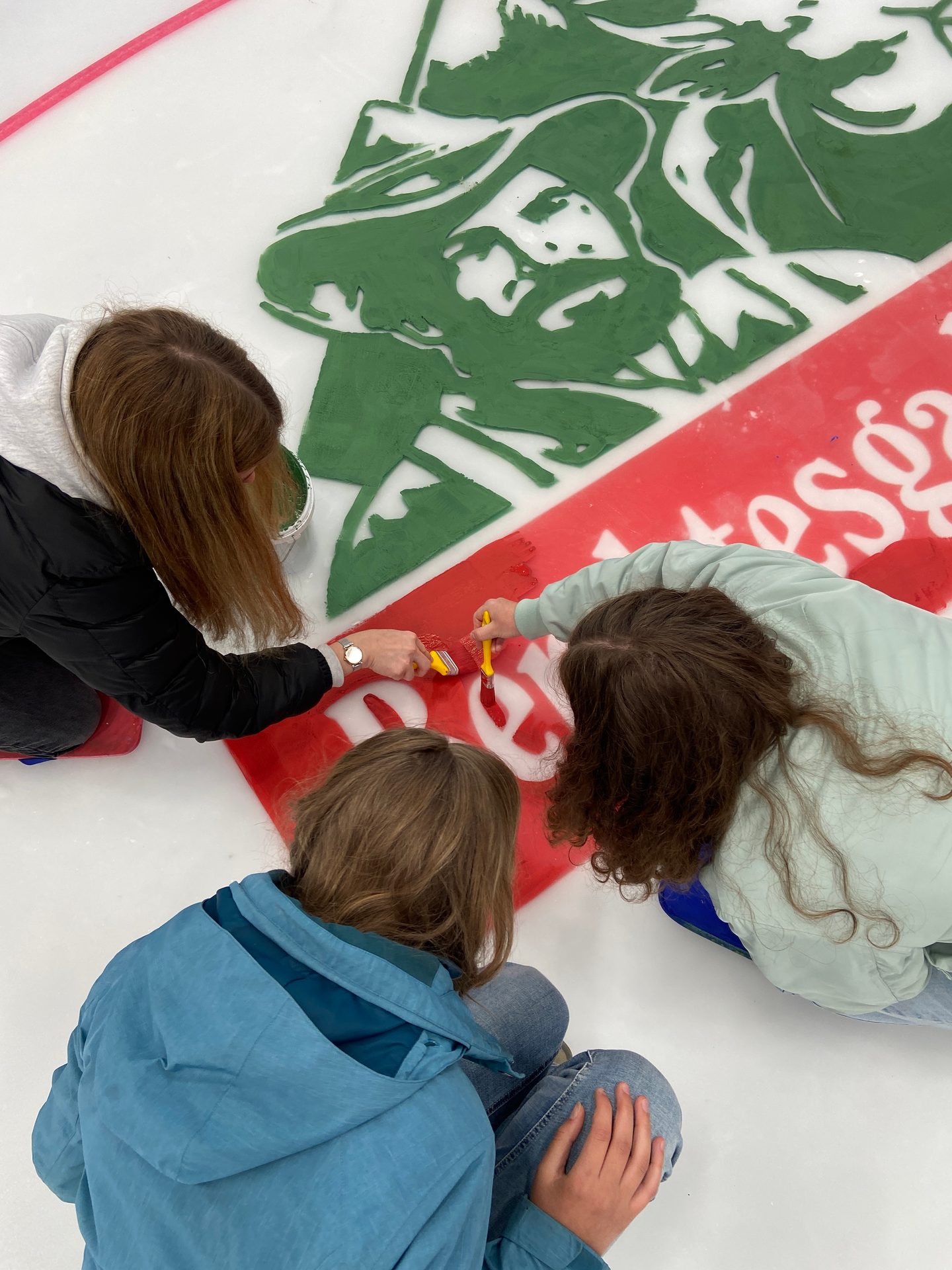 Three people paint a green and red stencil design on a white surface with brushes.
