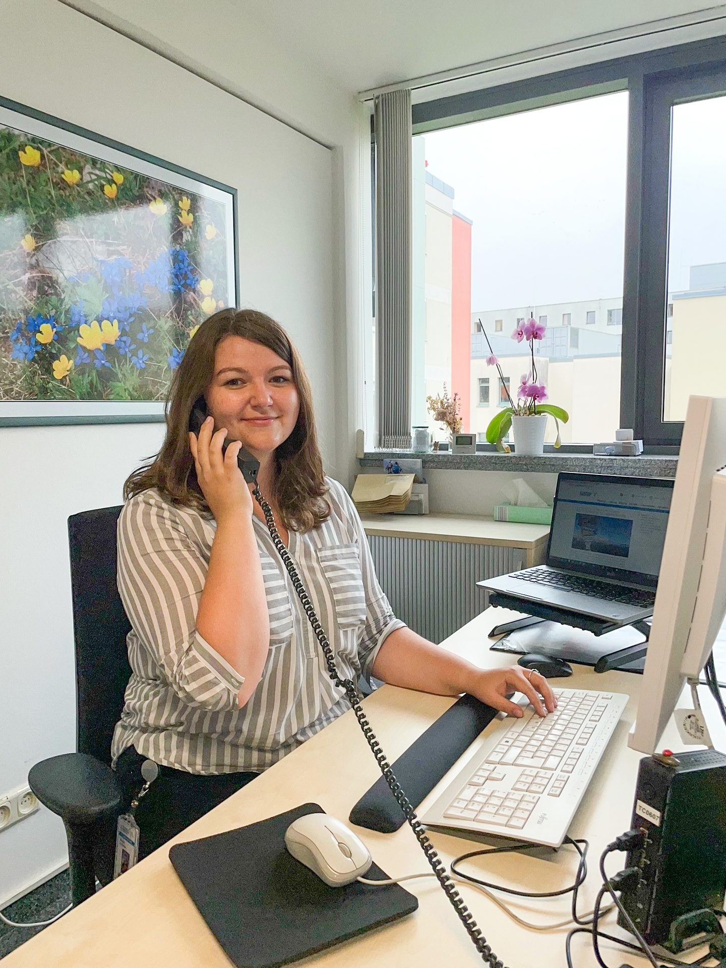 Smiling woman talks on a landline phone at an office desk with a computer and window.