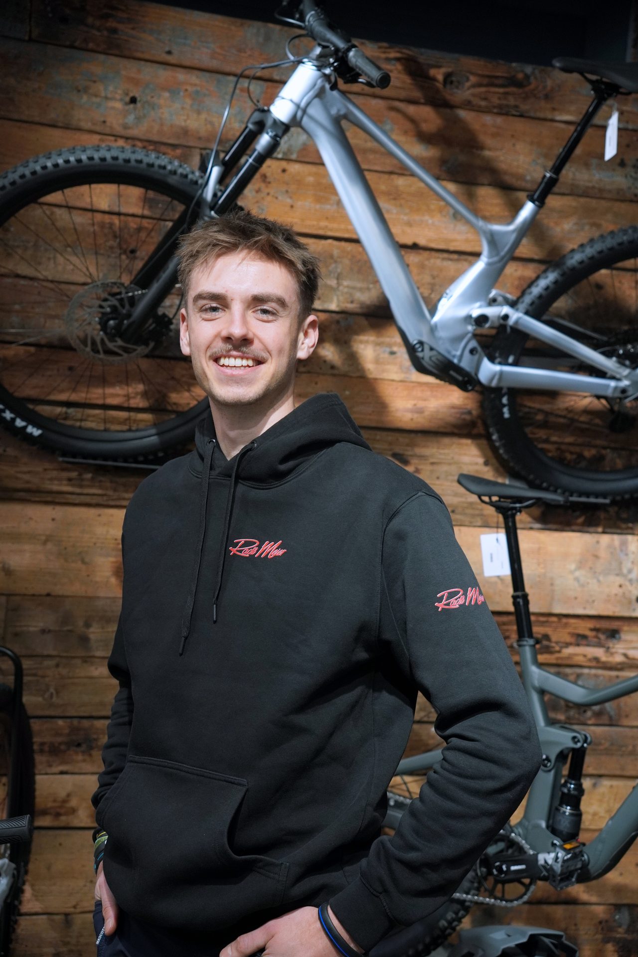 A smiling man in a black hoodie with a red logo stands in front of bicycles and a wooden wall.