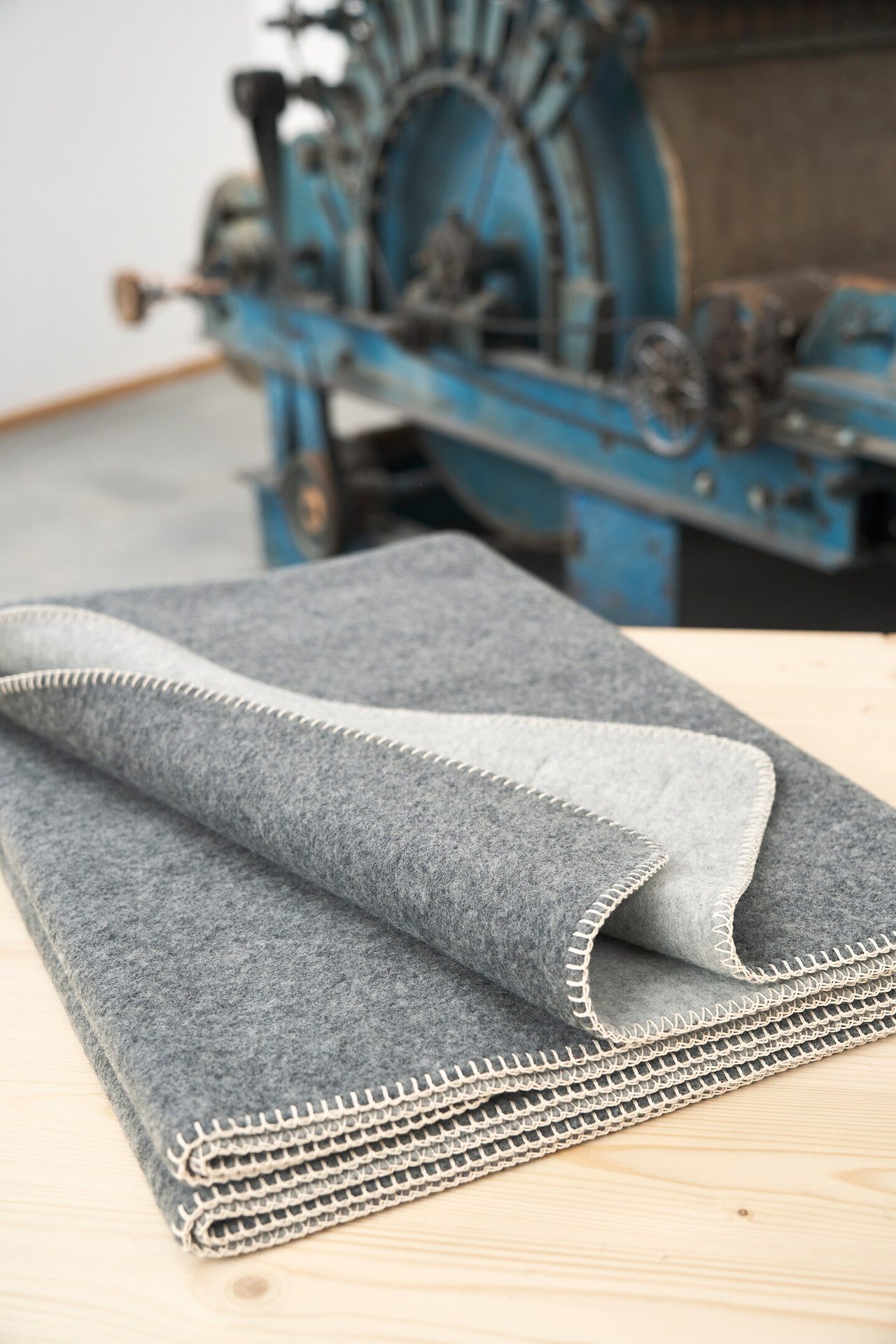 Folded grey felt blanket with white blanket stitch on wood, blurred blue factory machine in background.