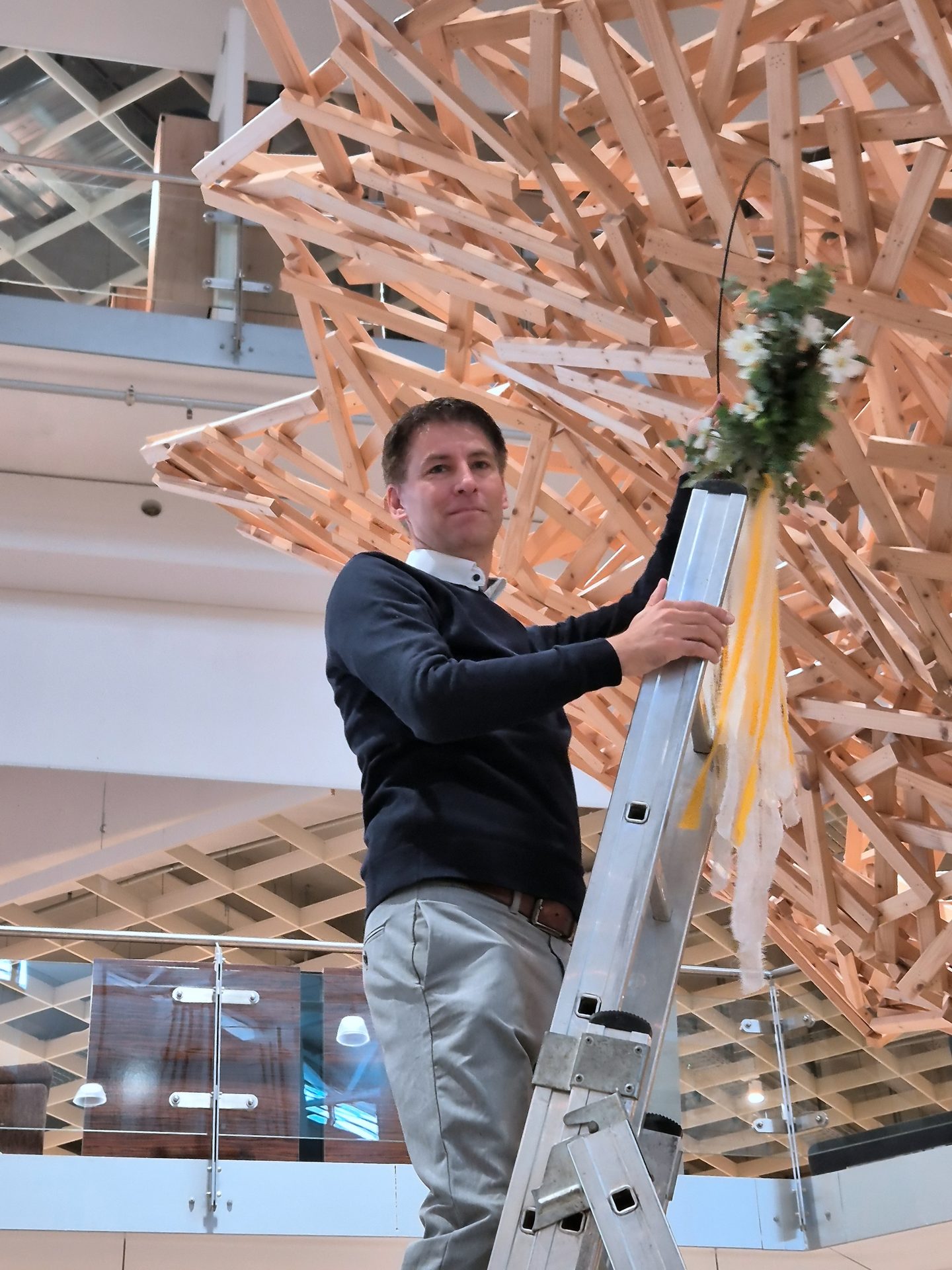 Man on ladder attaching flowers to a complex wooden art installation.