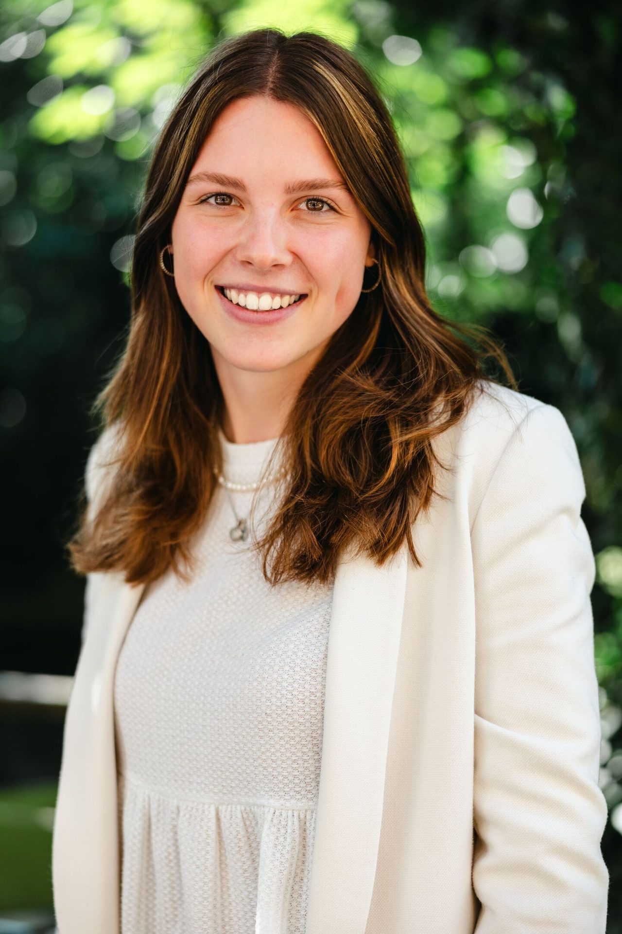 Smiling woman with long brown hair in white blazer against blurred green background.