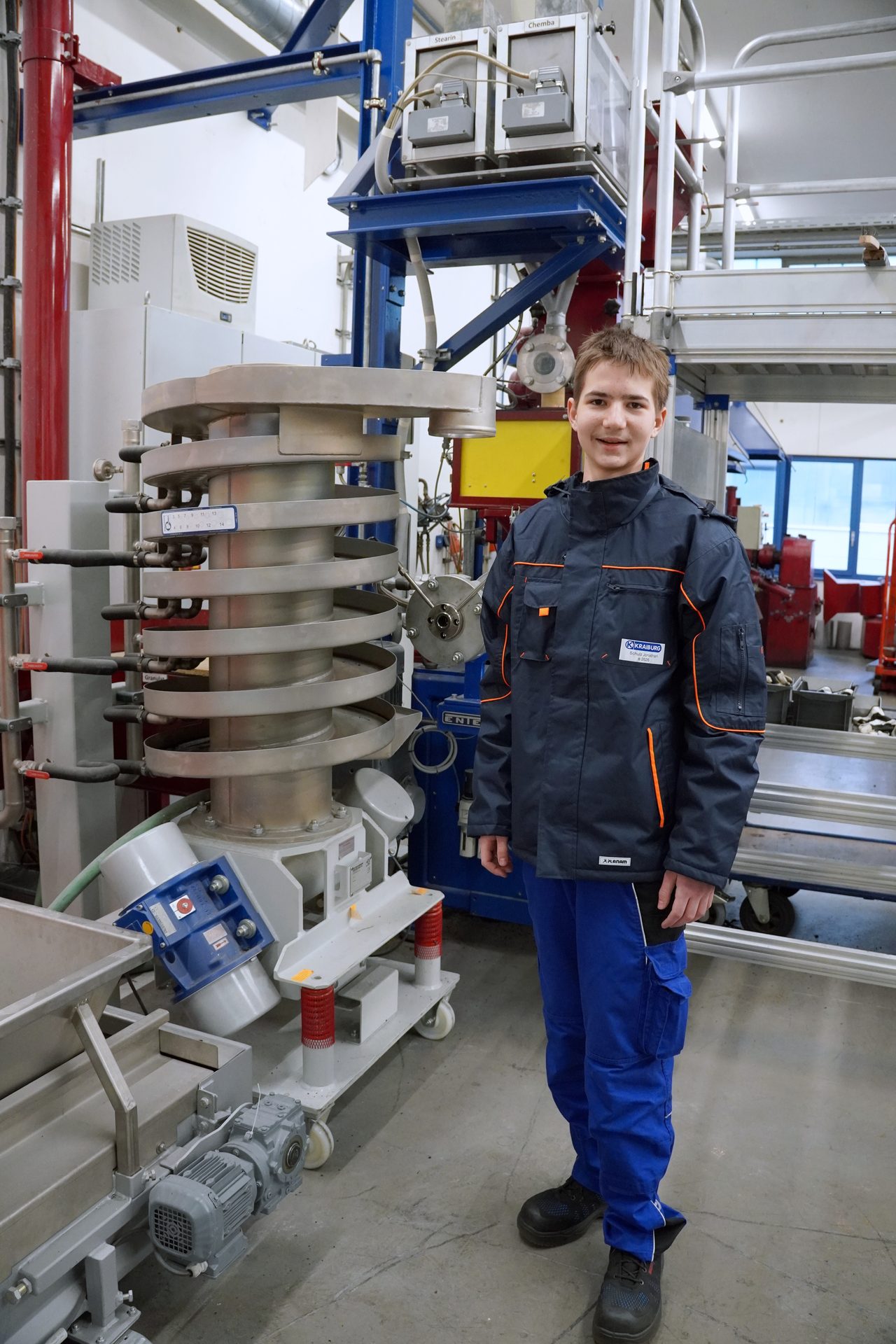Smiling young man in work uniform stands by industrial processing equipment.