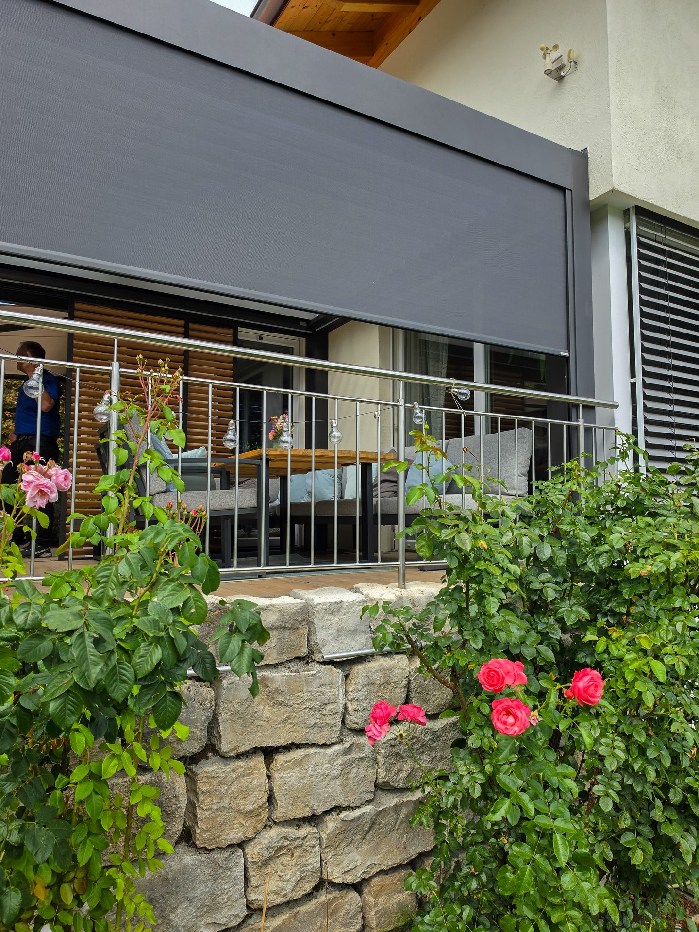 A grey retractable awning shades a balcony with outdoor furniture, seen above a stone wall and rose bushes.