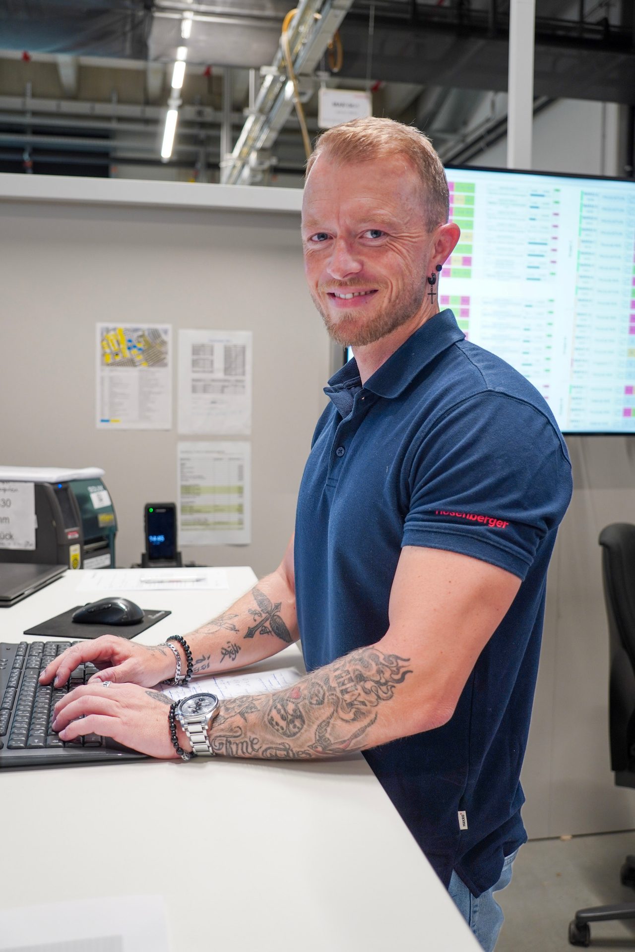 Smiling man with tattoos on his arms working at a computer in an industrial setting.