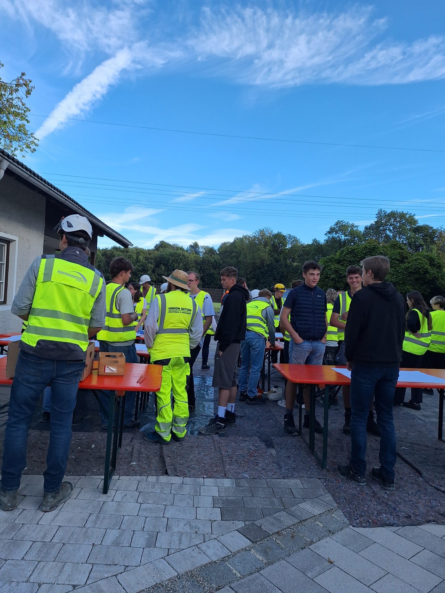 Young people in hi-vis vests gather around tables outdoors under a blue sky.