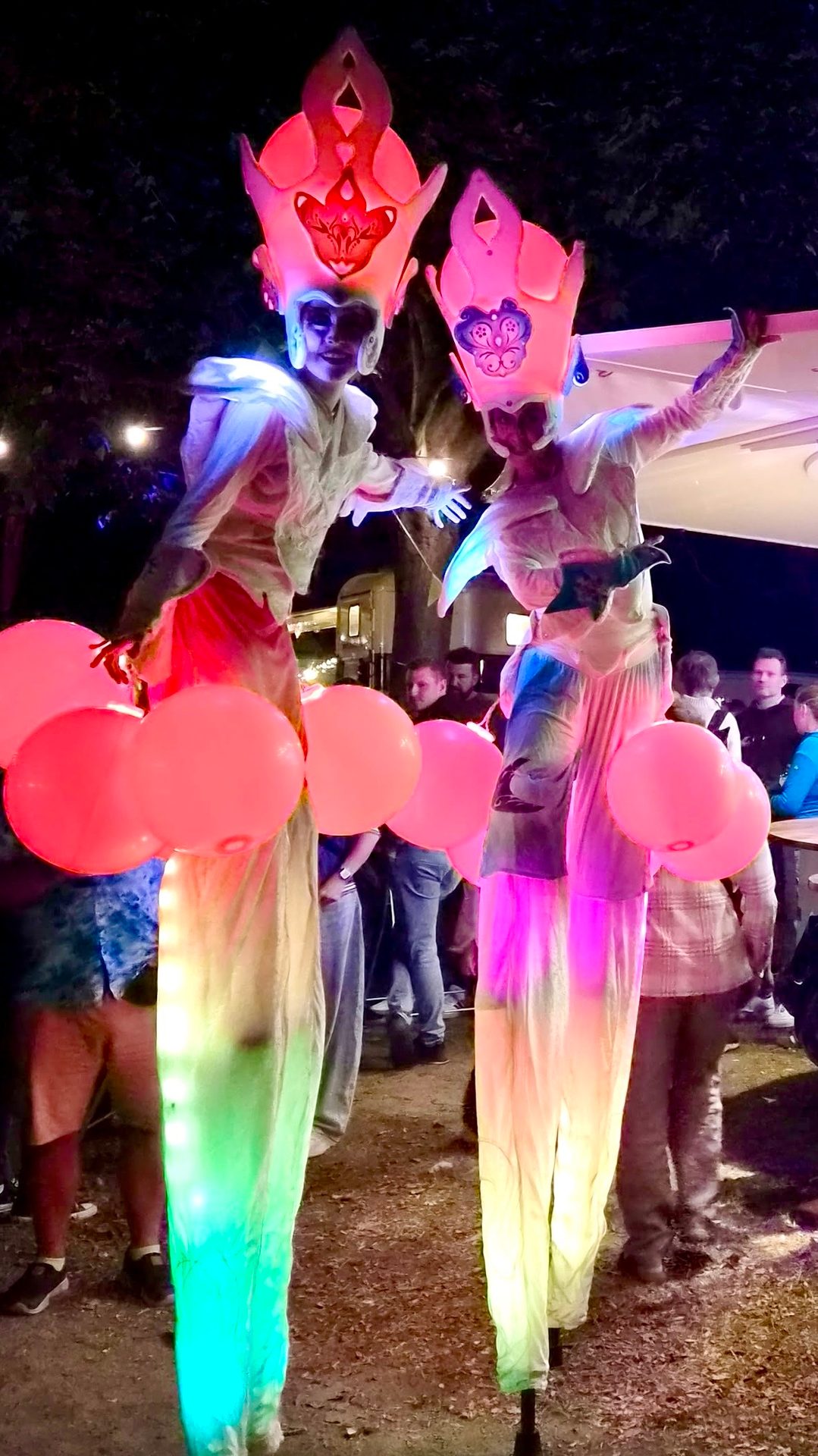 Two stilt walkers in glowing, elaborate costumes with illuminated hats and pink balloons perform at a night event.