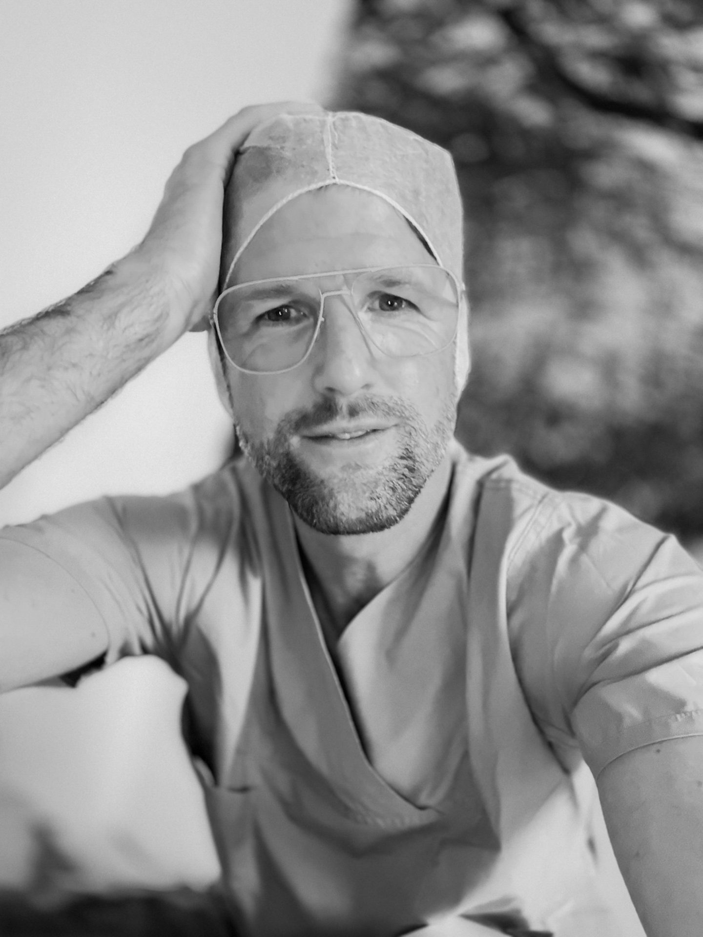 B&W portrait of a man in scrubs and a surgical cap, with glasses, looking at the camera.